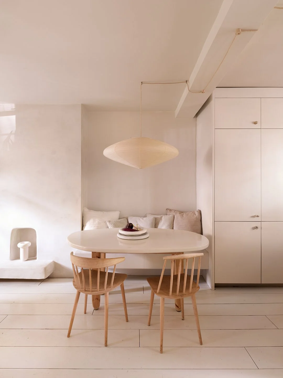Minimalist dining area with a round white table, two wooden chairs, a cushioned bench, and a hanging paper pendant light in neutral tones.