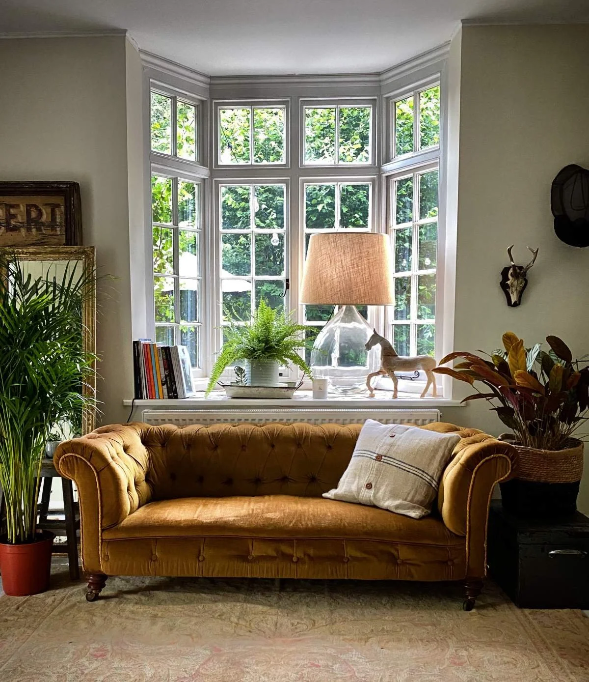 Living room with a mustard-colored vintage tufted sofa, a window with green trees outside, a lamp with a clear glass base and beige shade, a potted fern, a book arrangement, decorative horse figurines, and wall art including a deer skull and a hat.
