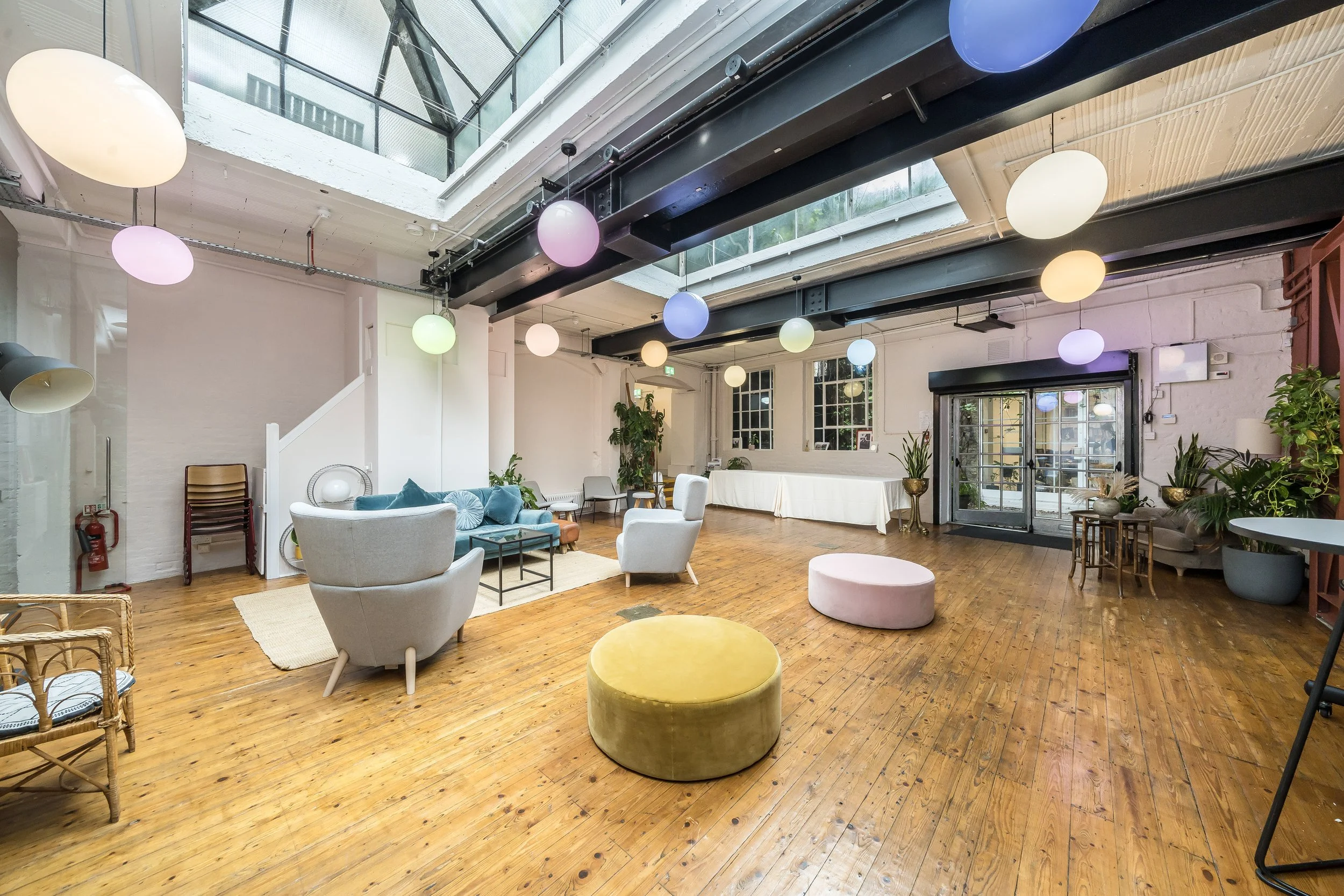 A spacious, modern lounge area with hardwood floors, white brick walls, and a glass ceiling. It features colorful hanging spherical lights, various couches, chairs, and ottomans, and numerous potted plants near the windows and door.