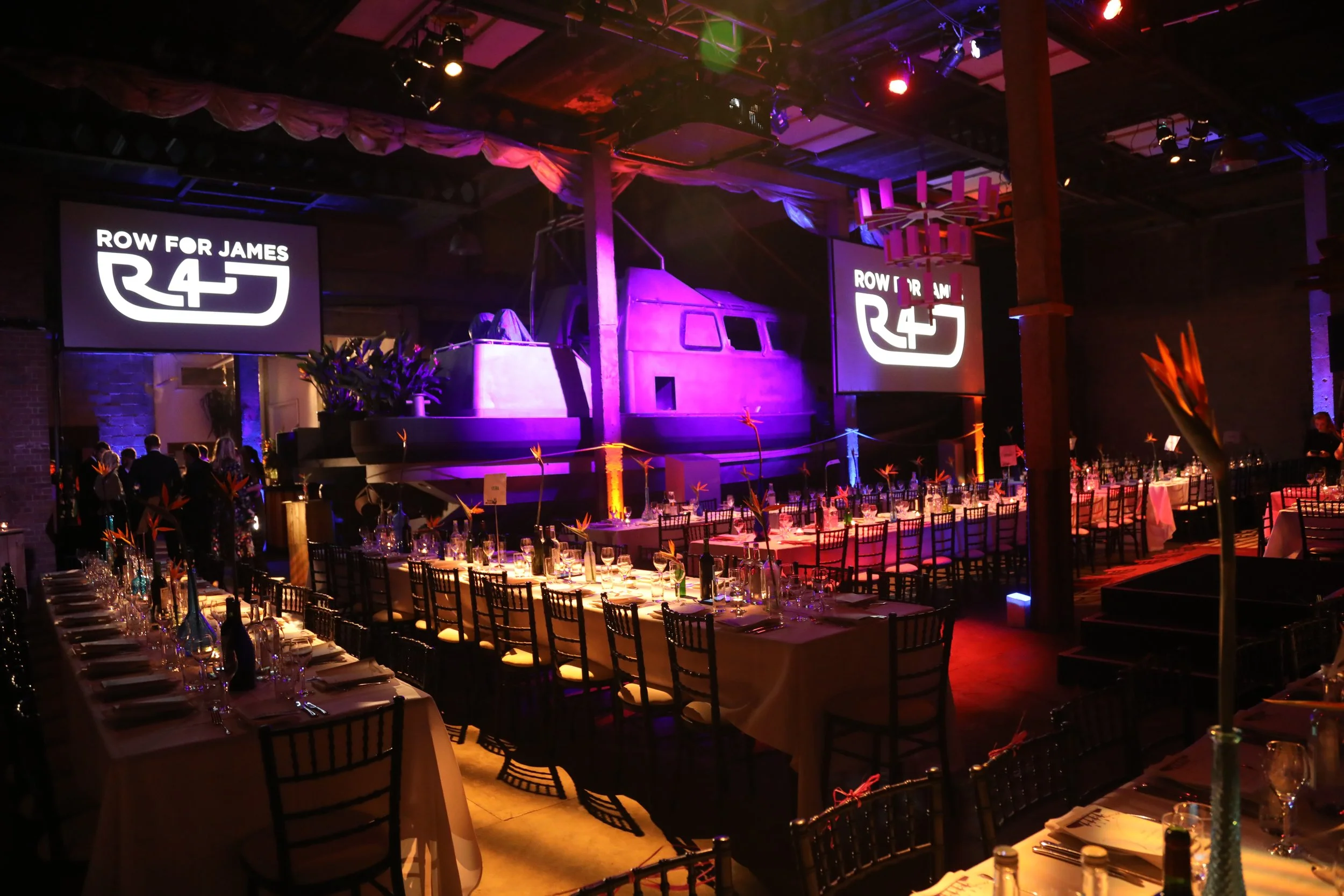 Indoor event space with long dining tables decorated with colorful vases and flowers, illuminated by purple and orange lighting, featuring a stage with a helicopter set piece and large screens displaying 'ROW FOR JAMES R4J' logo.