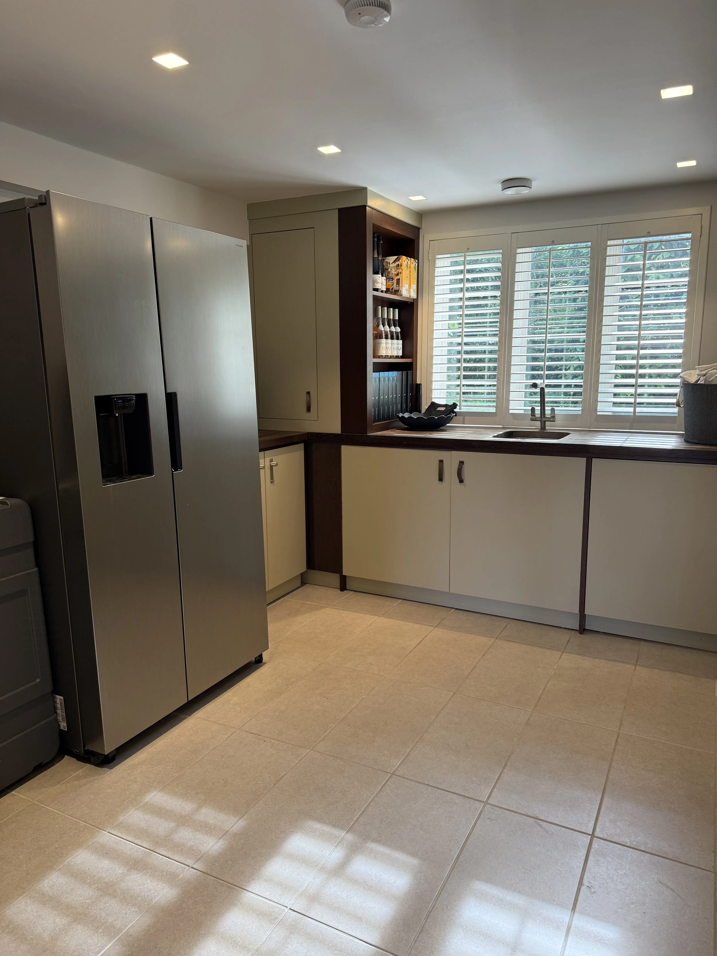 Modern kitchen with stainless steel refrigerator, beige cabinets, dark wood accents, large window with plantation shutters, and tiled floor.