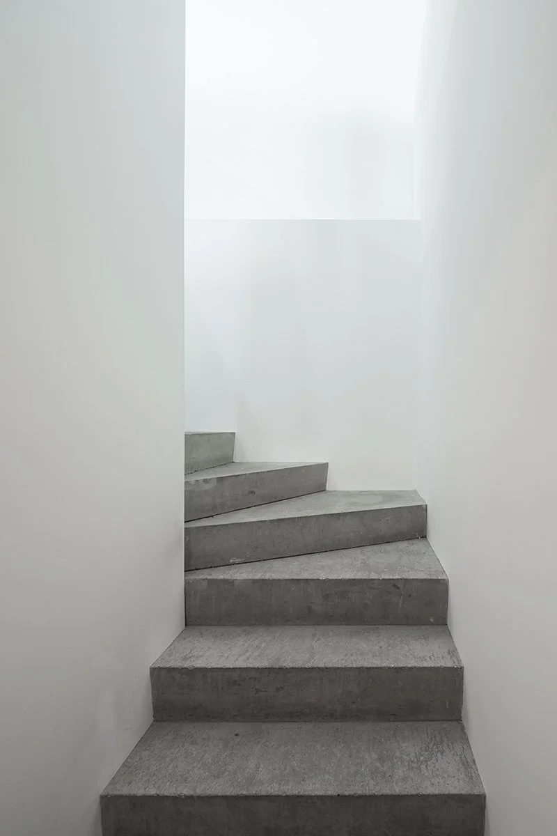 Concrete stairs in a minimalist white-walled interior with natural light coming from a window at the top.