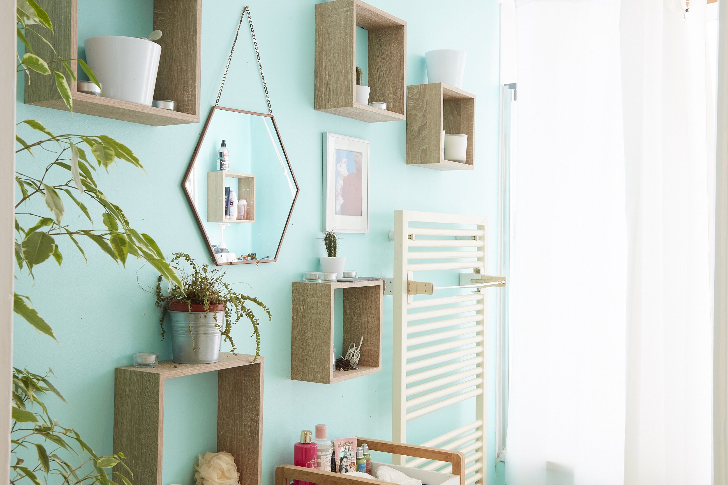 Decorative bathroom corner with a light blue wall, wooden shelves, a hanging mirror, potted plants, candles, and toiletries, illuminated by natural light through white curtains.