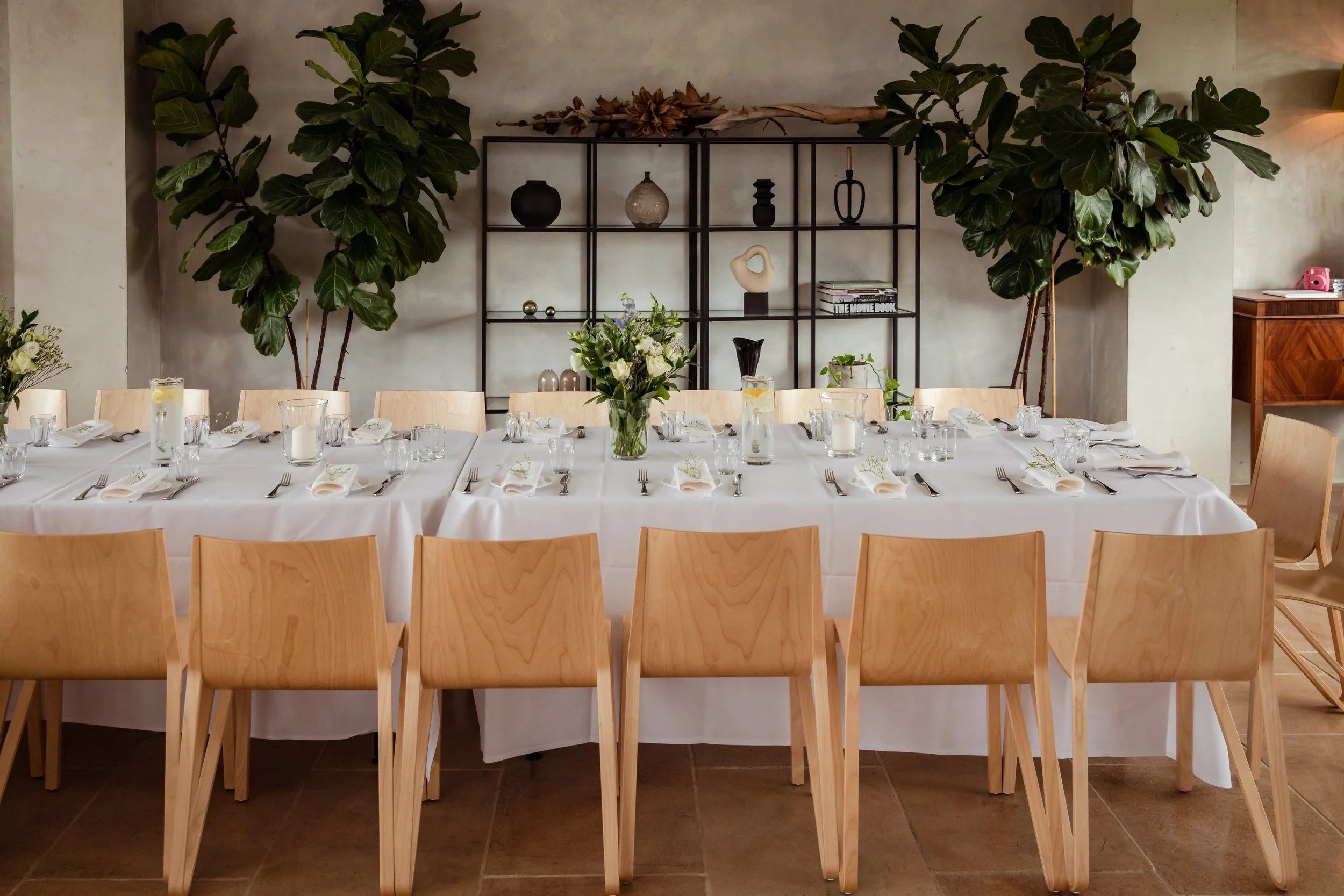 Dining table set for a special occasion with white tablecloth, glassware, and napkins, surrounded by wooden chairs, decorated with a floral centerpiece, in a modern room with large indoor plants and wall decor.