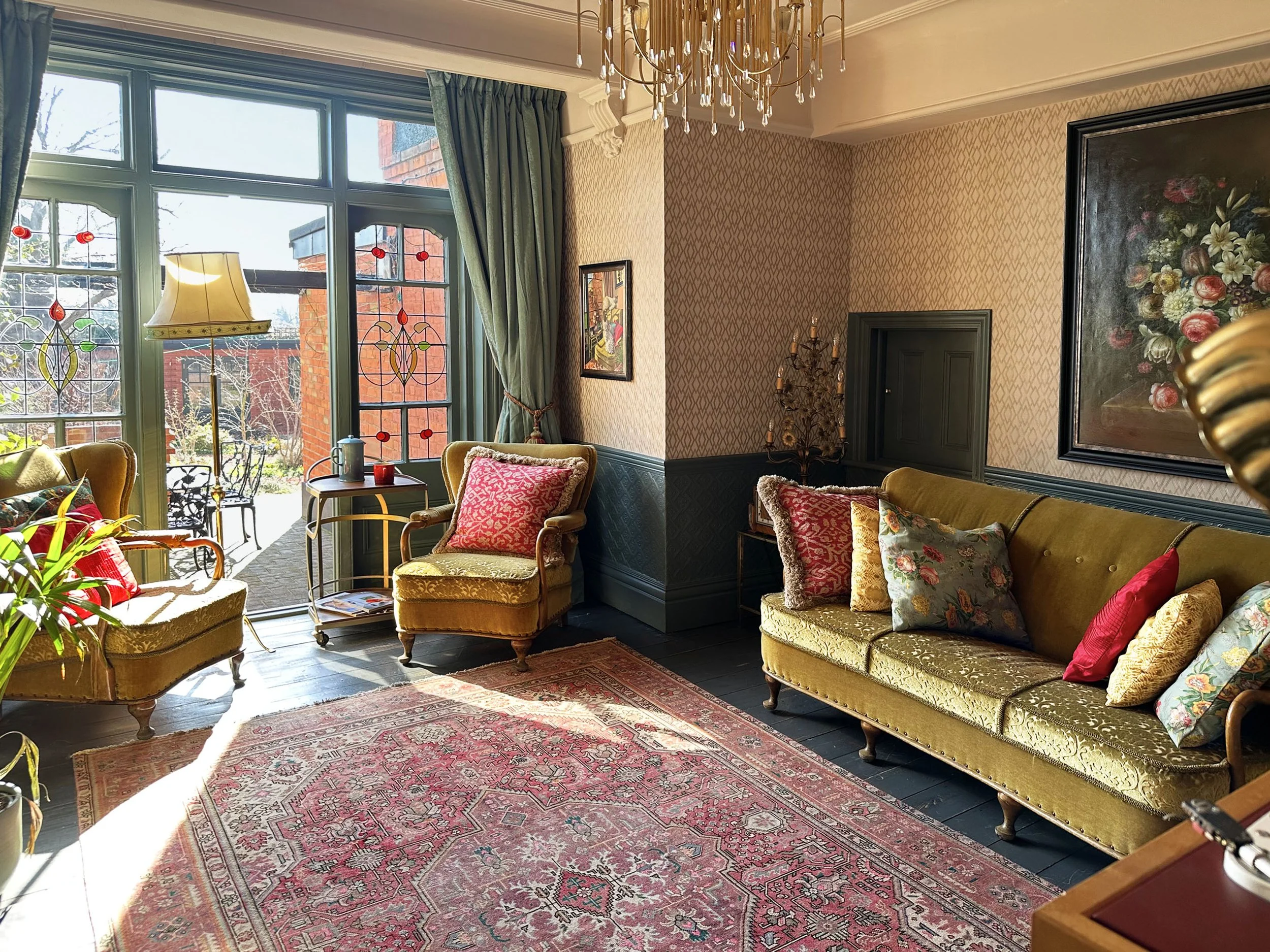 A cozy living room with vintage yellow-green upholstery sofas and armchairs, decorative pillows, a large floral painting on the wall, a chandelier hanging from the ceiling, and a glass door leading to an outdoor garden with brick buildings visible ou