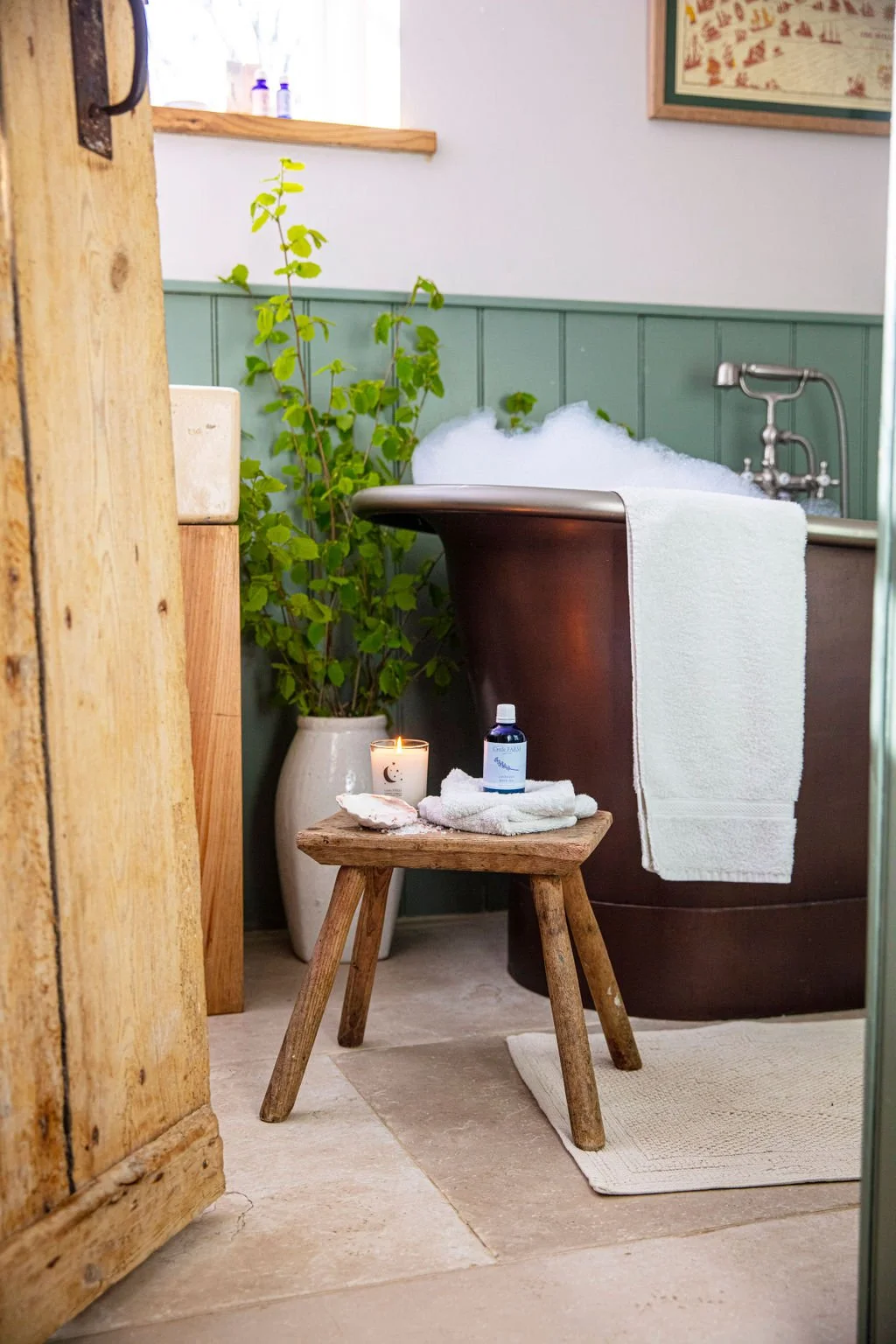 A vintage-style bathroom with a bathtub filled with bubbles, a small wooden stool holding a candle, a bottle of essential oil, a towel, and shells. There is a potted plant next to the bathtub, and a window with sunlight coming through.