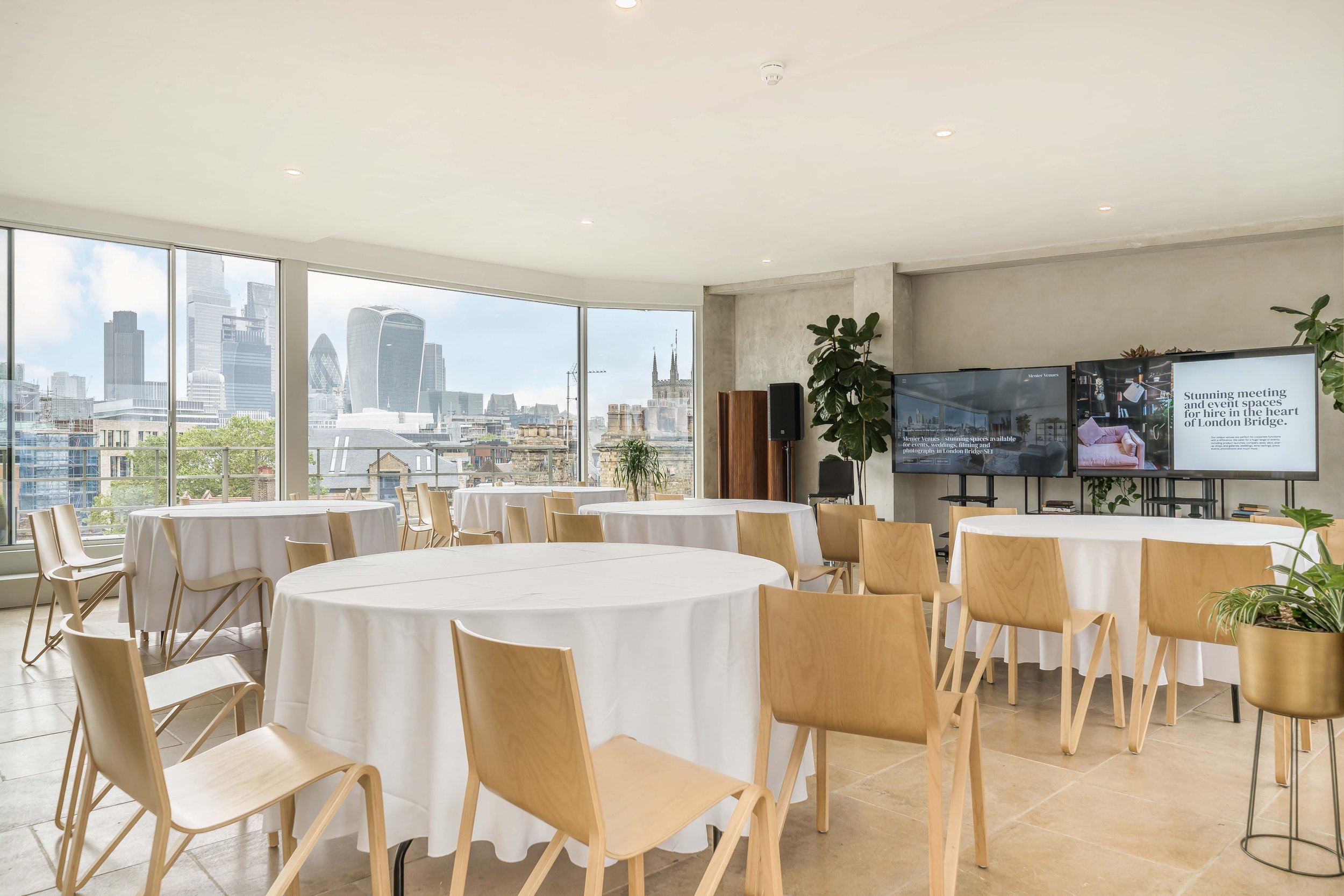 Conference room with round tables covered in white tablecloths, wooden chairs, large windows showing London city skyline, digital screens displaying meeting information, and potted plants.