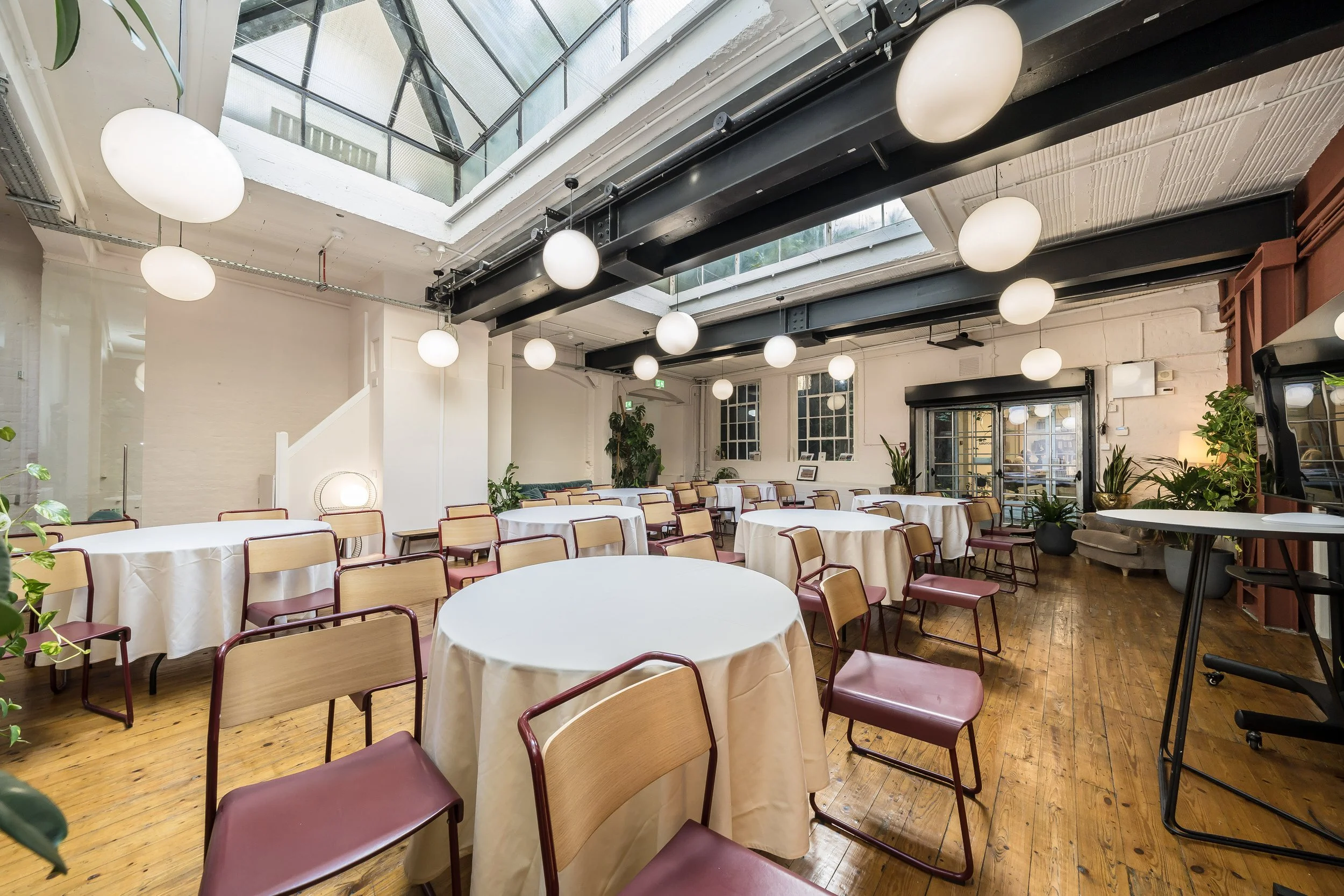 An empty event space or restaurant with round tables covered in white tablecloths and wooden chairs with maroon seats. The room has a large glass ceiling letting in natural light, and modern white globe pendant lights hanging from black beams. There 