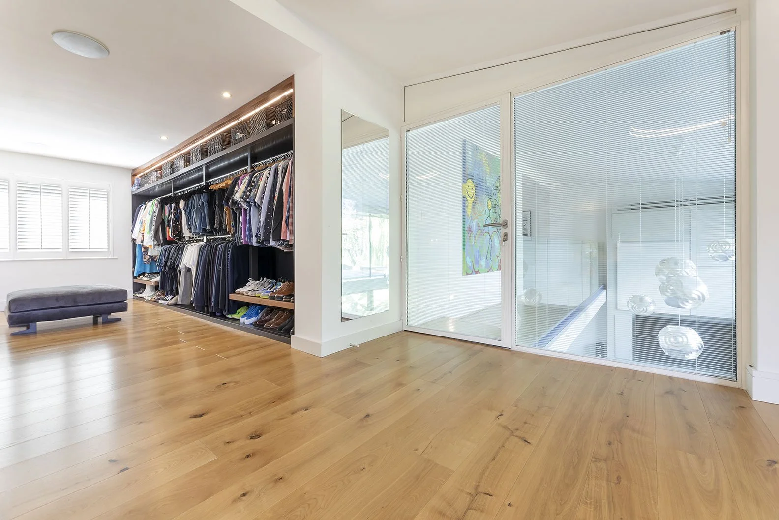 Modern walk-in closet with wooden floors, open clothing racks, and large glass doors with blinds, featuring a mirror and decorative items.