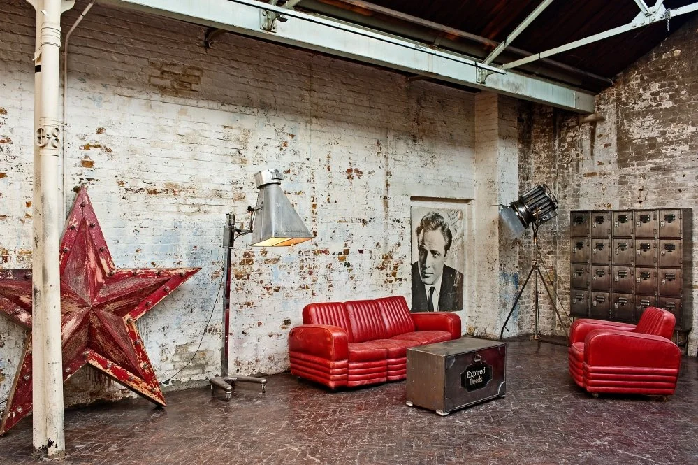 An industrial-style room with red vintage furniture, including a sofa and an armchair, a large distressed star decoration, a black and white portrait on the wall, lockers, and studio lighting.