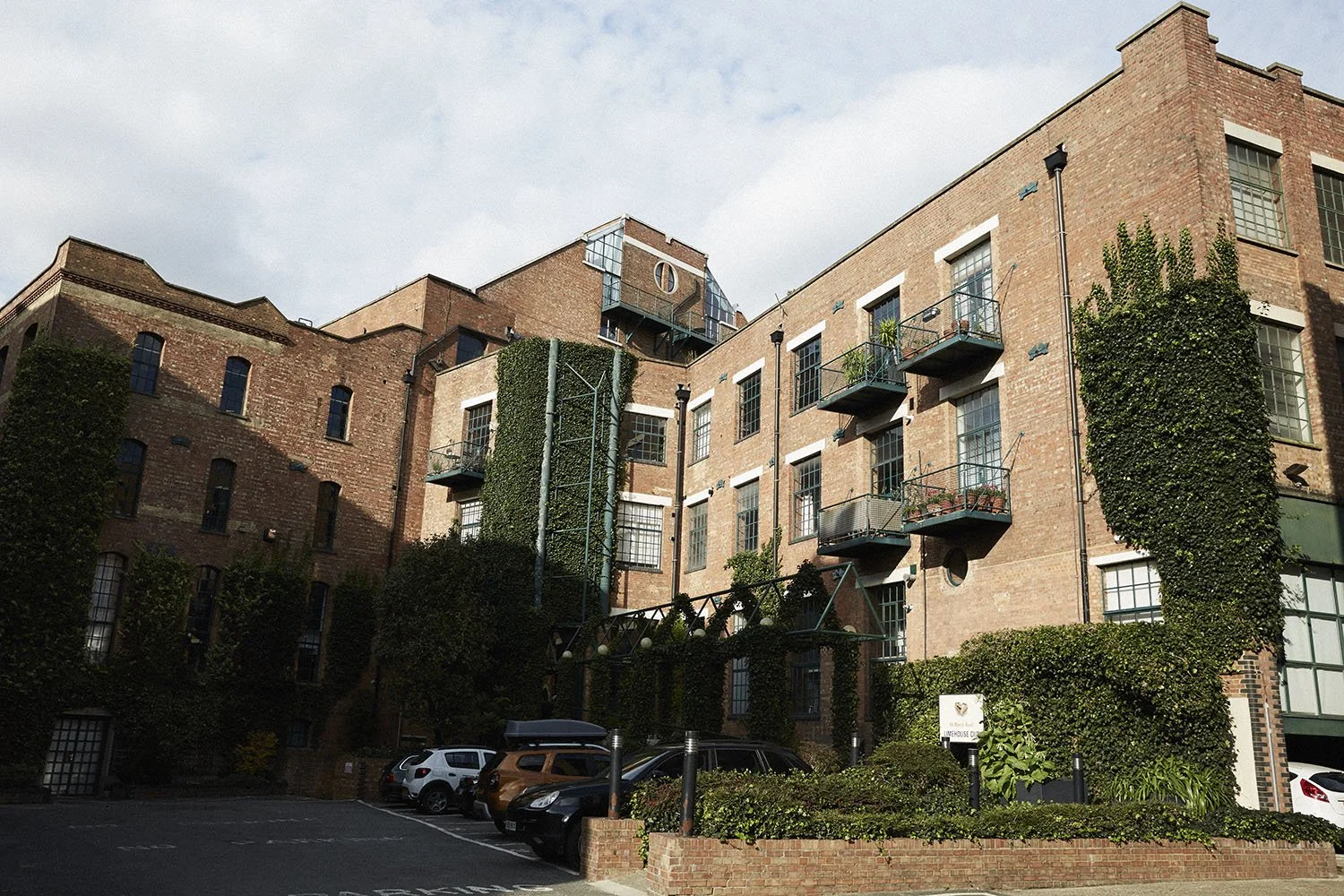 A multi-story red brick residential building with small balconies, surrounded by greenery and parking spaces in the foreground.