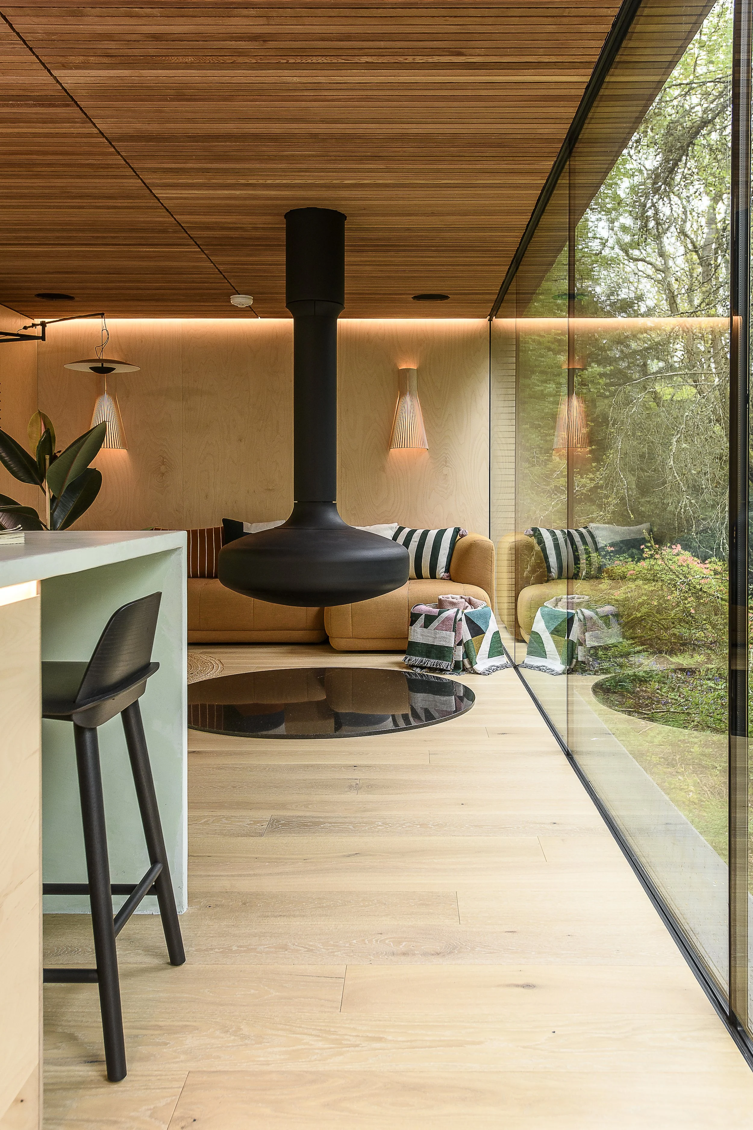 Modern living room with wooden ceiling and walls, large glass window overlooking garden, yellow sofa with striped cushions, black hanging fireplace, black coffee table, and stylish wall sconces.