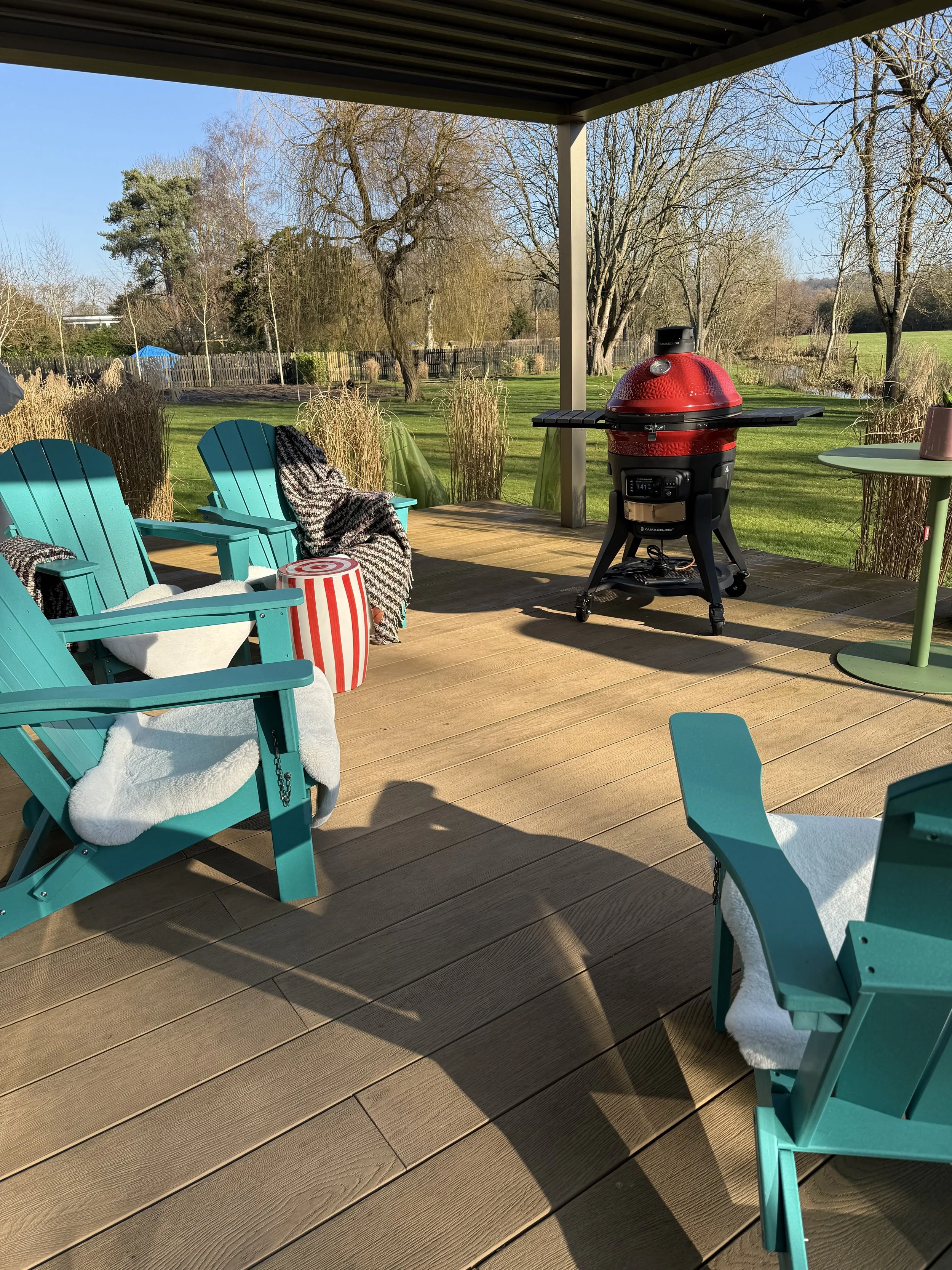 A backyard patio with four teal Adirondack chairs, one draped with a blanket, and a small round table. There is a red ceramic grill on a stand. The patio has a wooden deck and is covered by an overhang. In the background, there is a grassy yard with 