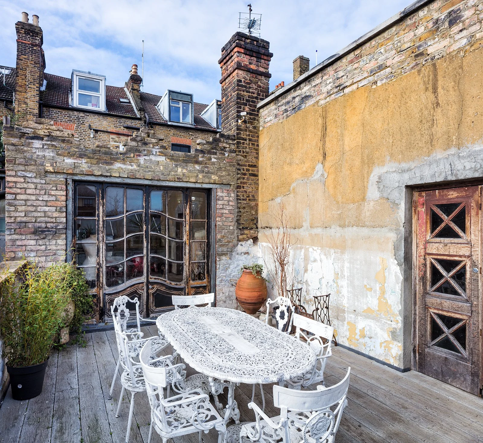 Urban rooftop terrace with a white ornate metal dining table and chairs, potted plants, a large clay pot, and a weathered wooden door, surrounded by brick and stucco walls under a cloudy sky.