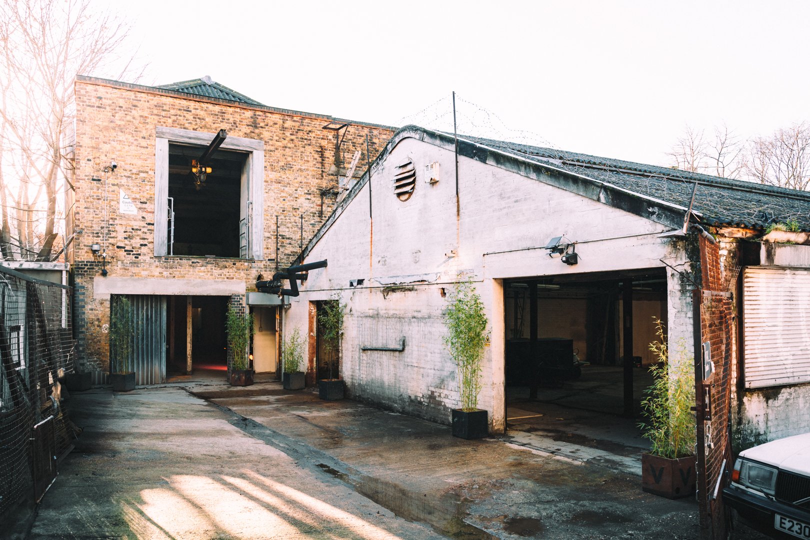 An urban alleyway with old brick and concrete buildings, some plants in pots near the entrances, a water pipe, security cameras, and a partially open roll-up garage door on the right.