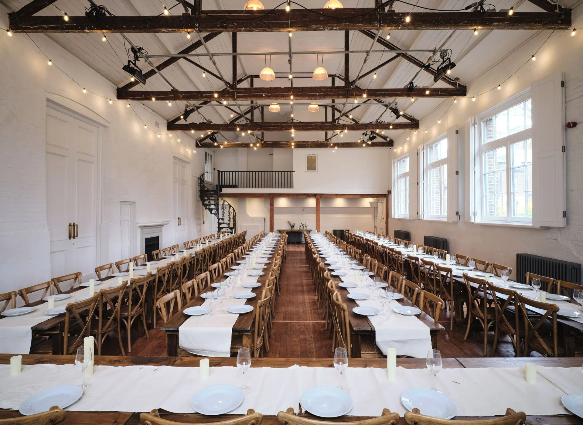 An interior banquet hall set up for an event with long wooden tables covered with white tablecloths, white plates, wine glasses, candles, and wooden chairs. The room has high ceilings with exposed wooden beams, string lights, large windows on the sid