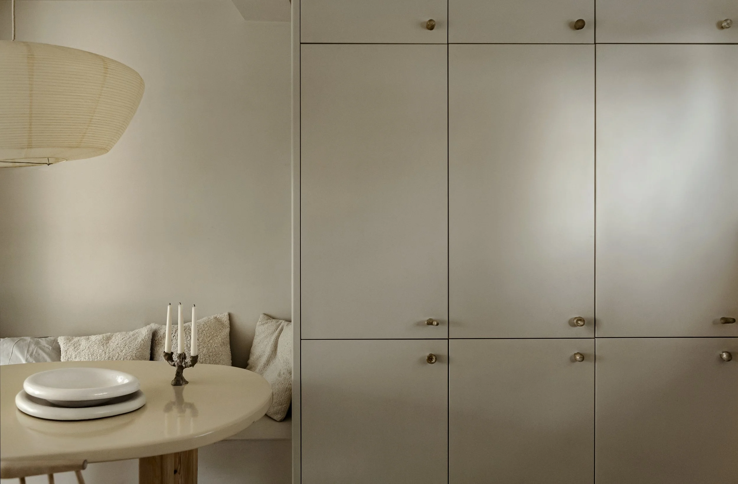 Dining area with a round table, stacked white plates, a candelabra, an upholstered bench with pillows, and a large beige cabinet in the background.