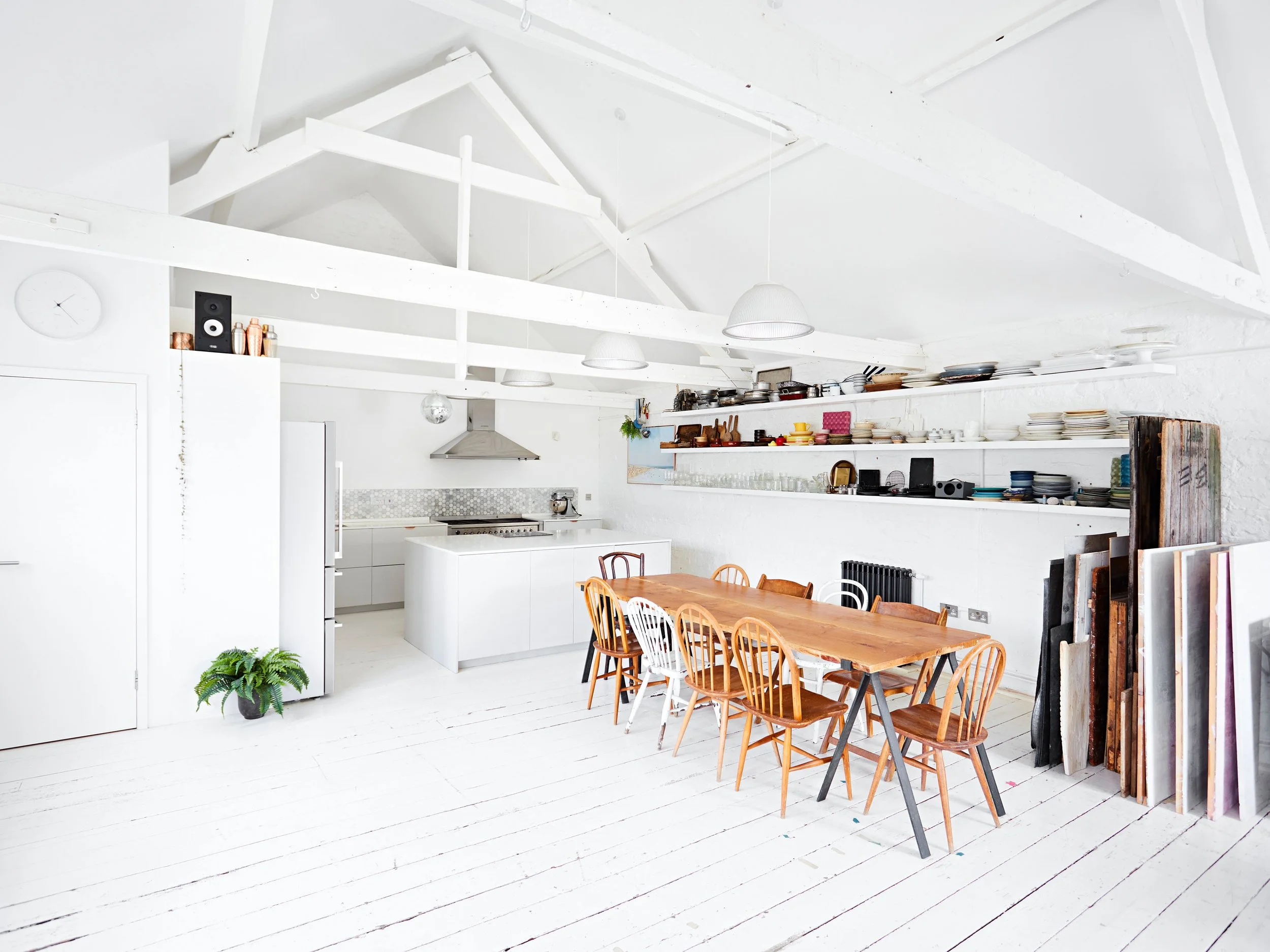 Bright, white kitchen and dining area with wooden table, mixed chairs, open shelves with dishes, and high cabin ceiling with exposed beams.