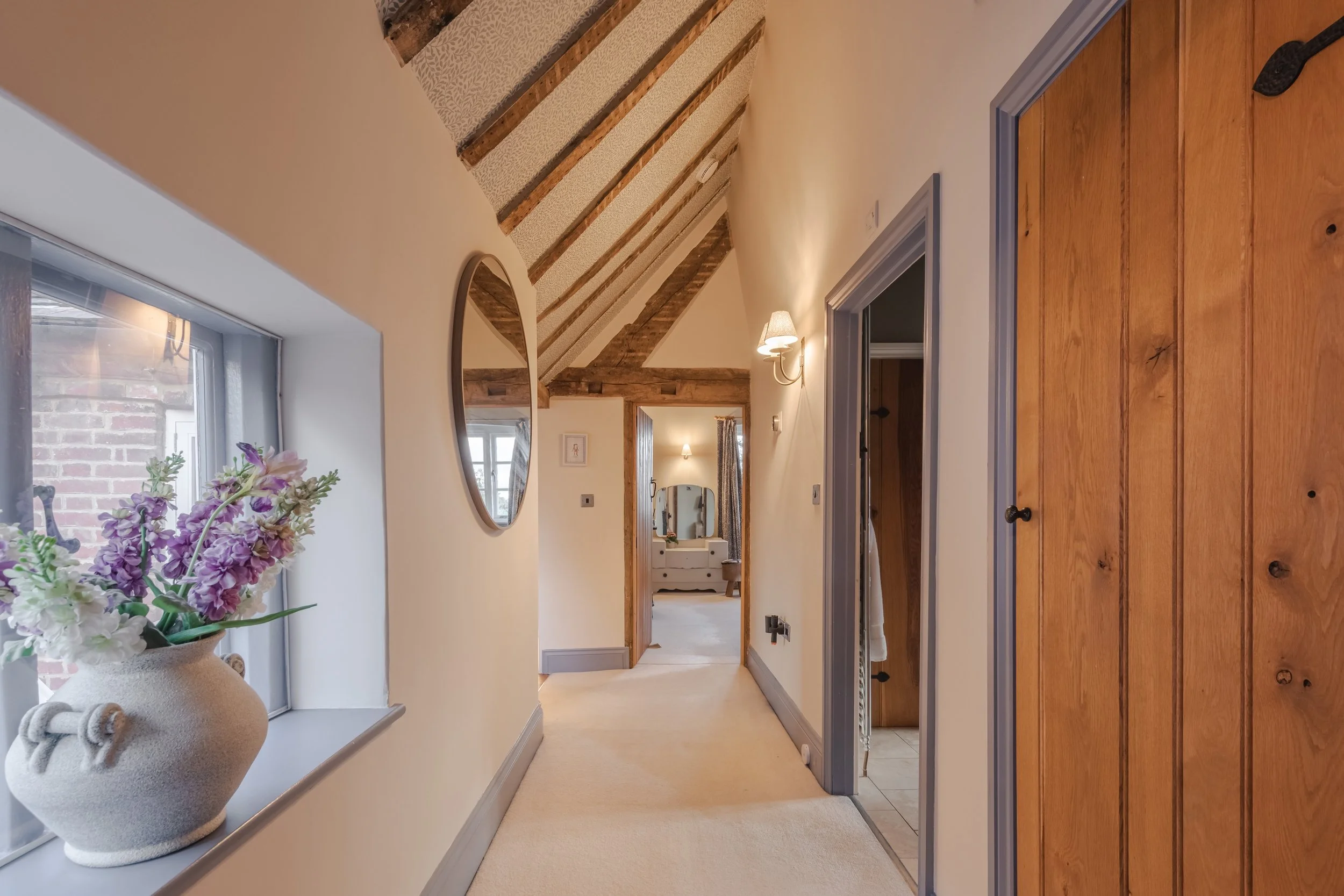 A hallway with cream-colored walls, a beige carpet, and wooden ceiling beams, featuring a window with a vase of purple and white flowers and framed mirrors.