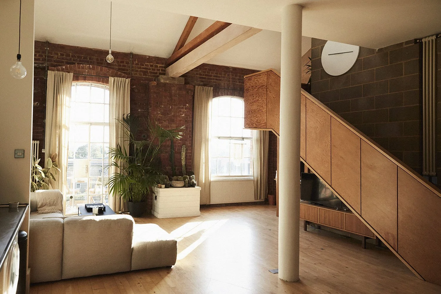 Living room with exposed brick walls, large windows with cream curtains, a white sectional sofa, potted plants, wooden staircase, and a television.