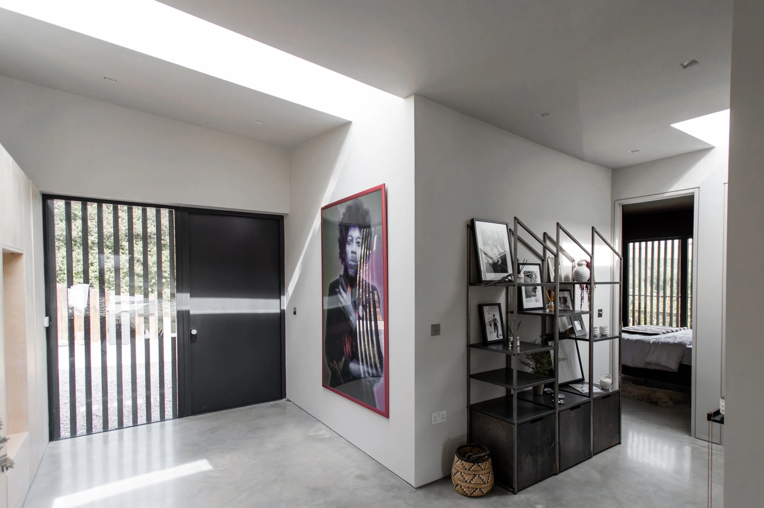 Modern interior hallway with a large black door, a portrait of a woman with an afro on the wall, a black metal shelf with framed photos and decor, and a doorway leading to a bedroom with a bed and window with blinds.