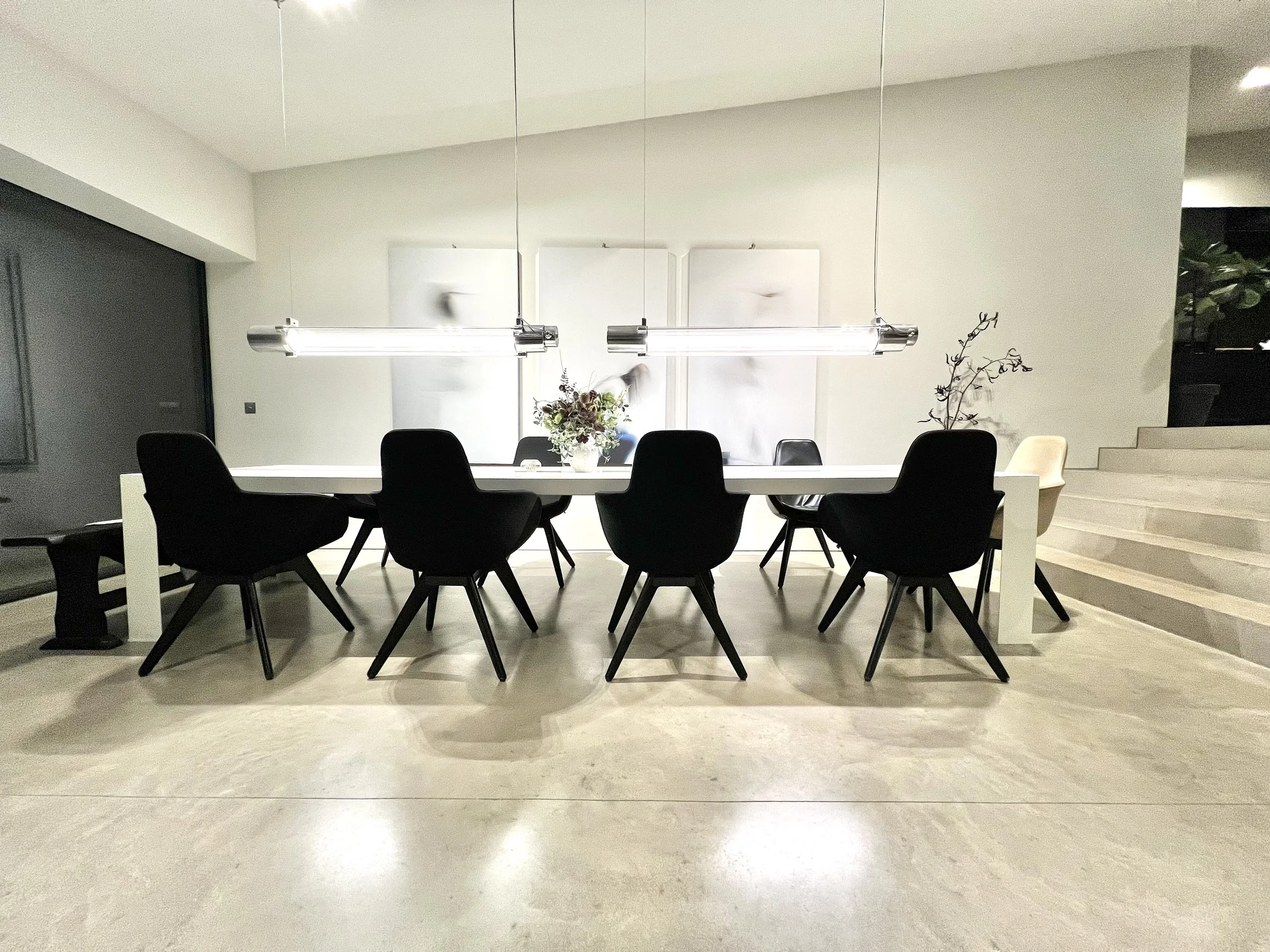 Modern dining room with a long white table, eight black chairs, a light fixture above, and minimalistic decor including a vase with flowers and a branch in a pot.