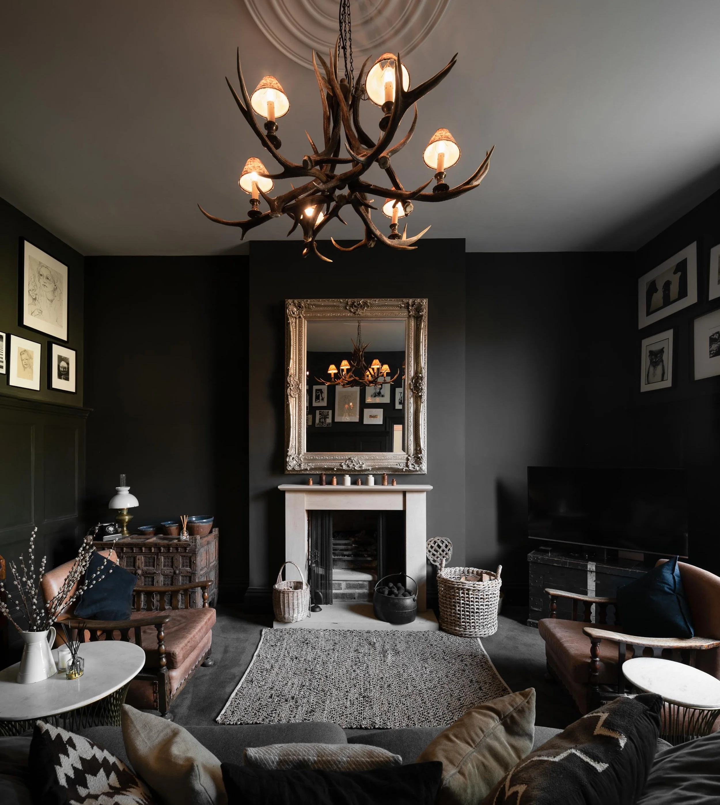 Living room with black walls, a white fireplace with a large ornate mirror above it, and a chandelier made of antlers with candle-shaped bulbs hanging from the ceiling. Furniture includes two armchairs, a sofa with pillows, and side tables. Framed ar