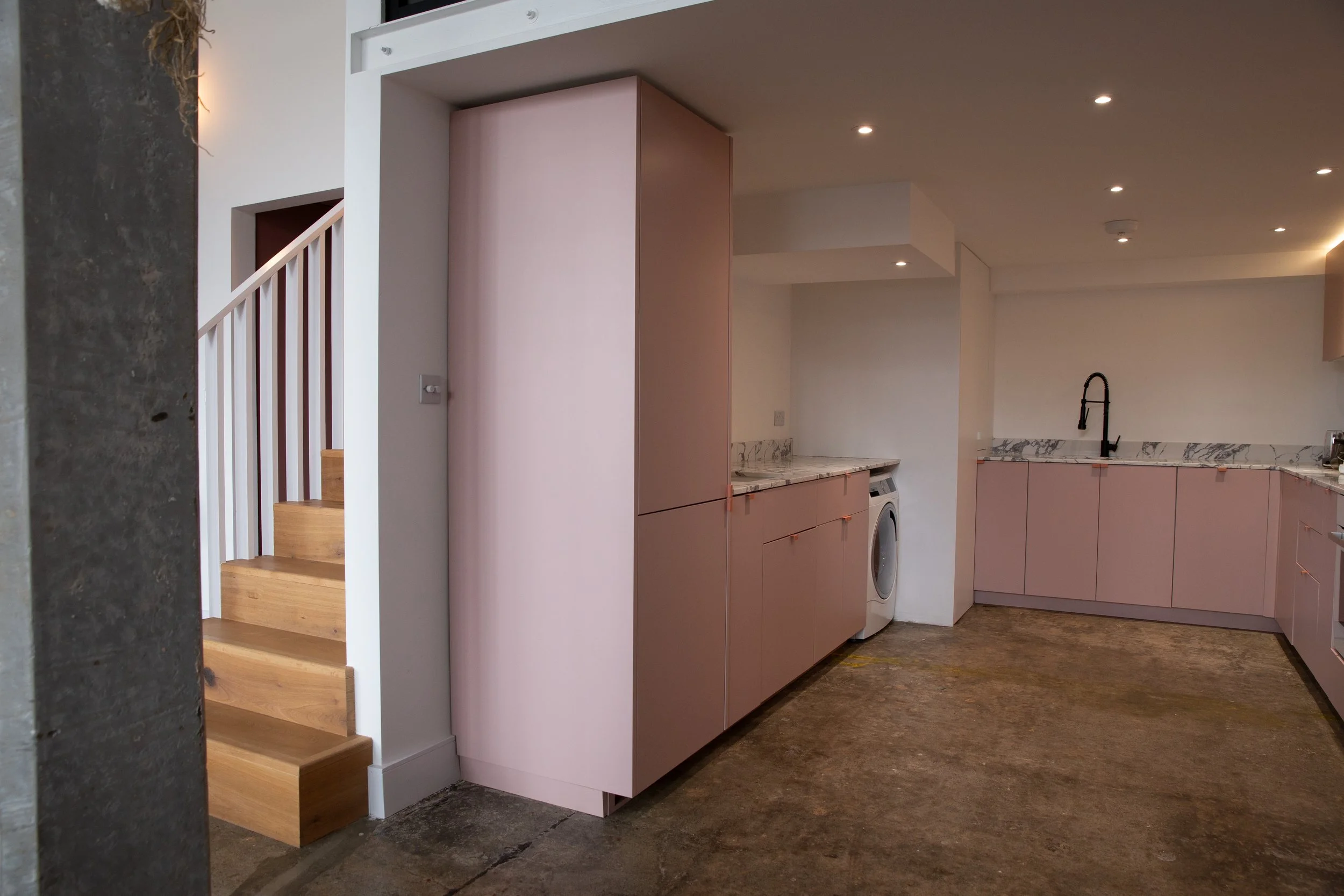 Modern kitchen with pink cabinets, marble countertops, and a black faucet, adjacent to a staircase with wooden steps and white railing.