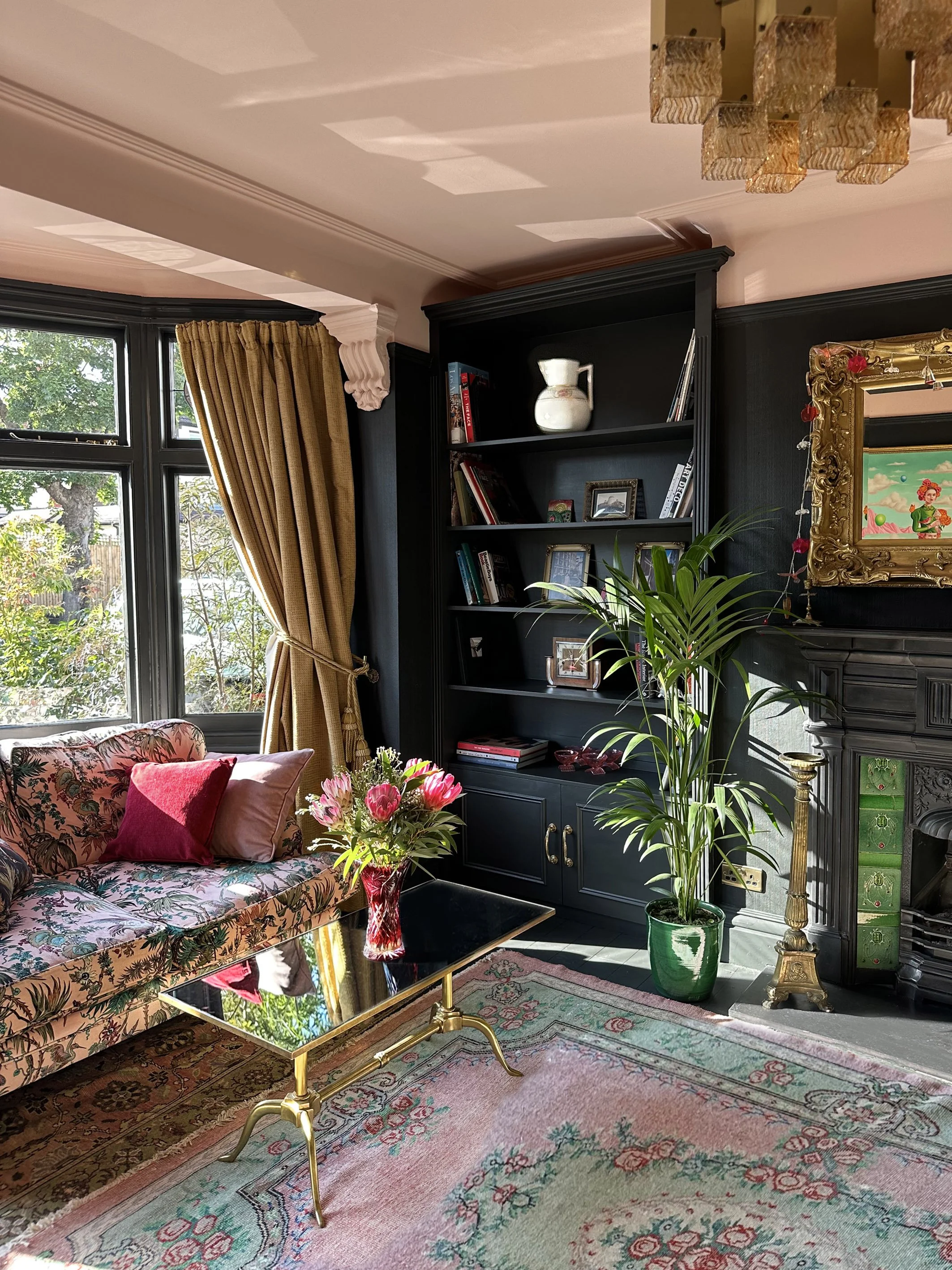 Living room with floral couch, gold and black furniture, black shelves, large window with golden curtains, pink flowers on a table, and green potted plants.