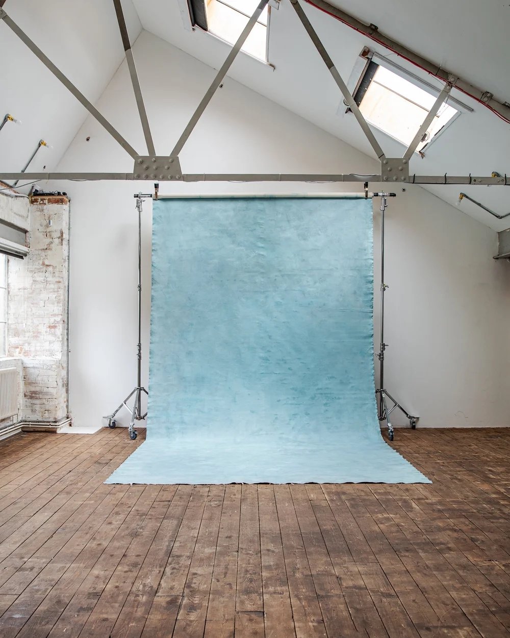Photo of an empty studio with a blue backdrop on a stand, wooden floor, white walls, and windows in the ceiling.