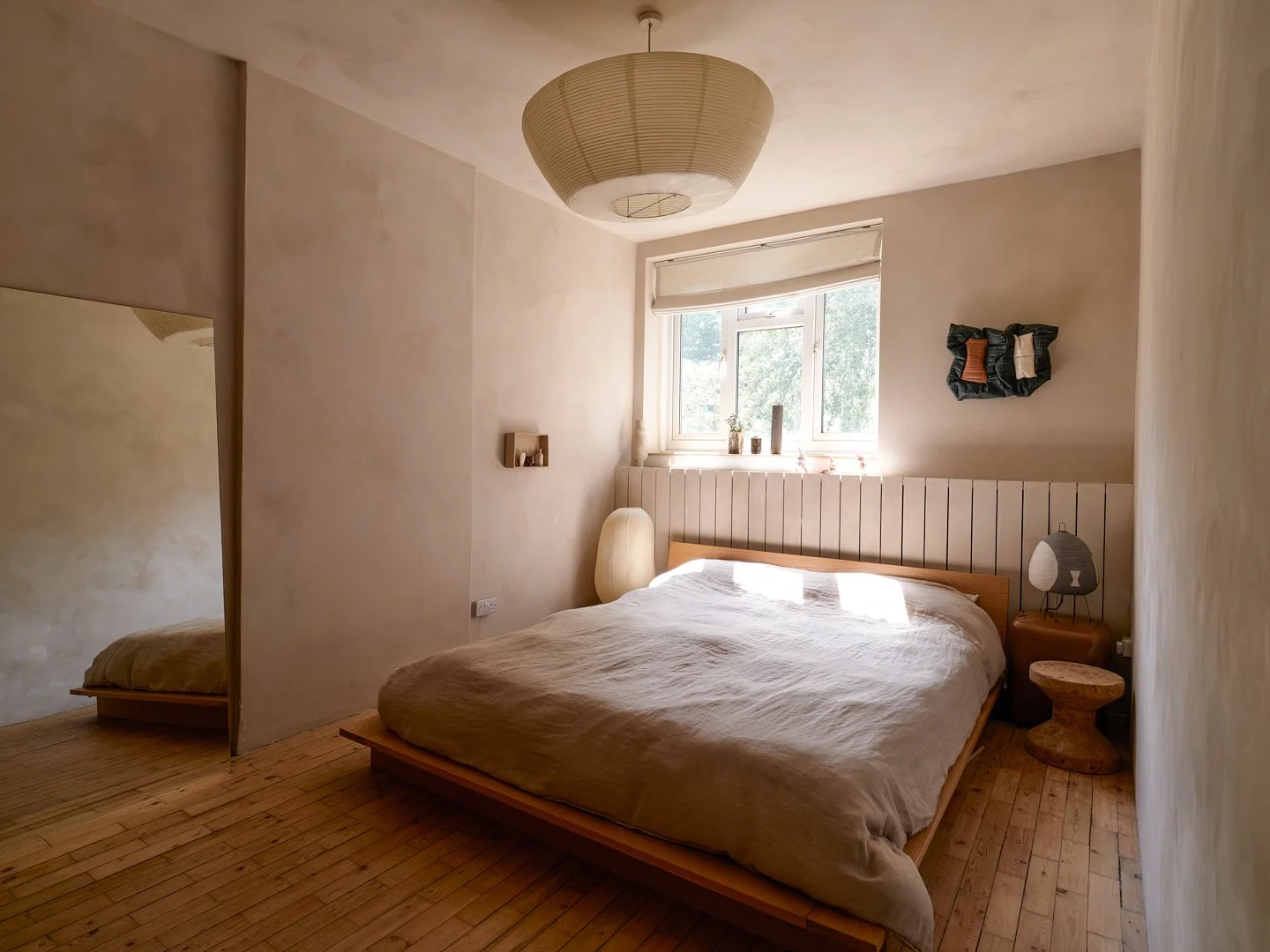 Minimalist bedroom with a wooden bed, beige bedding, a bedside table with a lamp, a window with sunlight, and natural decor elements.