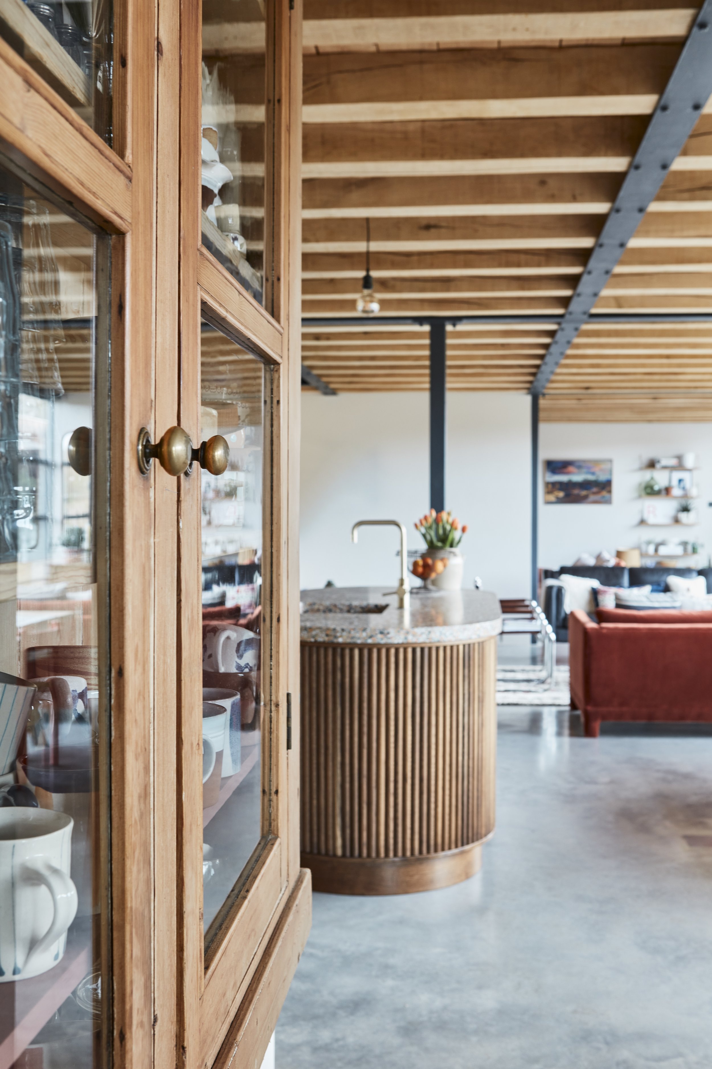A wooden cabinet with glass doors displaying mugs inside. The background shows a modern open living space with a kitchen island, a hanging light bulb, a vase with flowers, a dining table, and a living area with sofas and shelves.