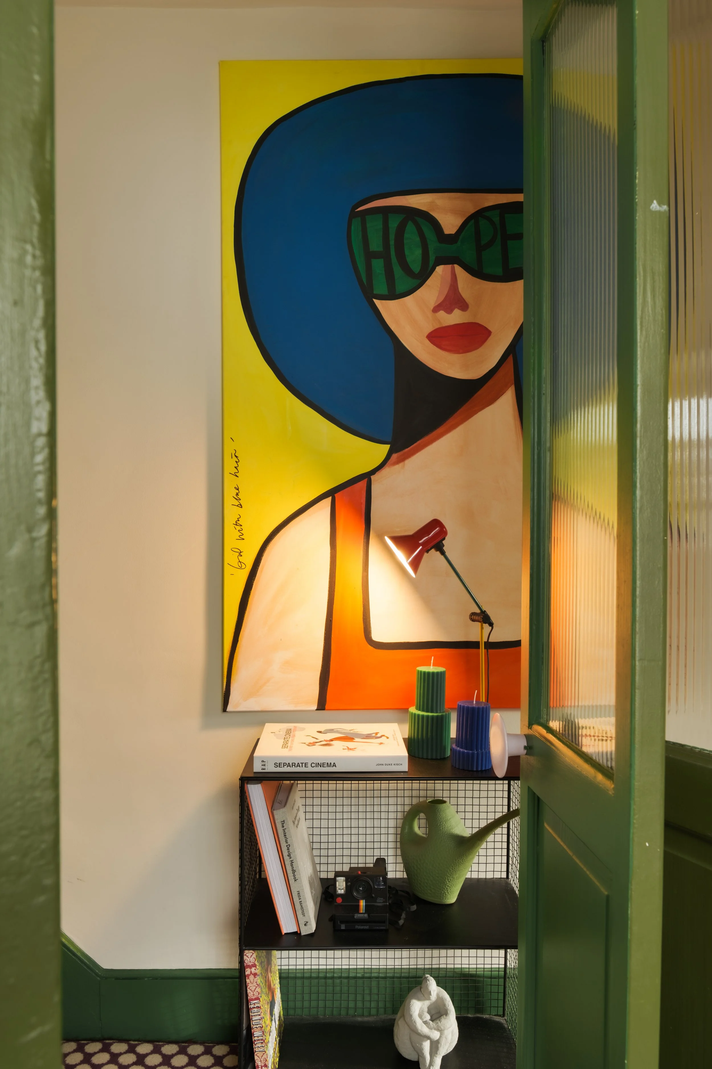 A colorful living space corner with a large painting of a woman wearing sunglasses and a blue hat, a small black metal shelf holding books, a vintage camera, a green watering can, decorative candles, a small lamp, and a white sculpture, with green an