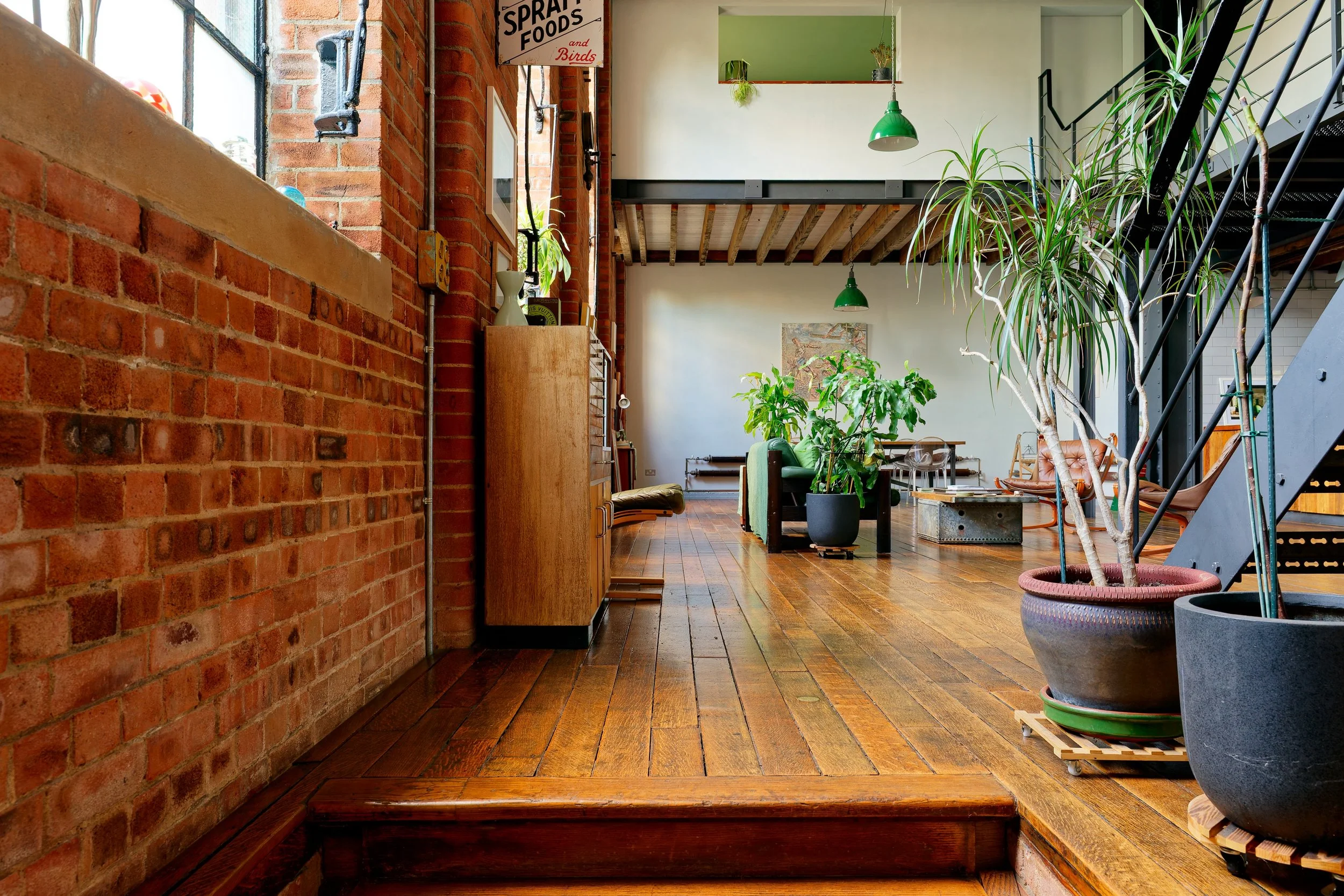Interior view of a modern loft-style apartment with brick walls, wooden flooring, and an abundance of potted plants. There are industrial-style green pendant lights, a staircase, and furniture including a sofa and chairs.