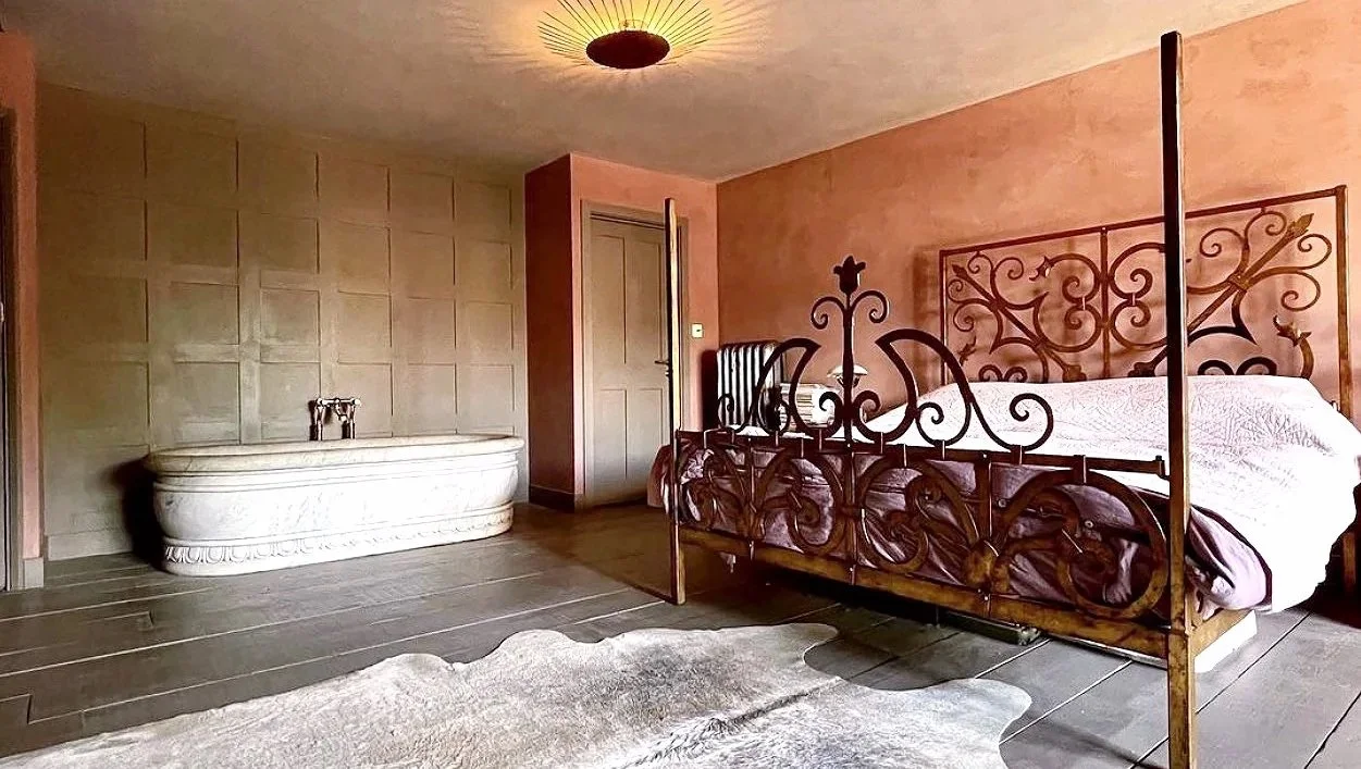 A bedroom with an ornate metal bed frame, a round chandelier on the ceiling, a textured wall with a closed door, a bathtub in the corner, and a cowhide rug on wooden floorboards.