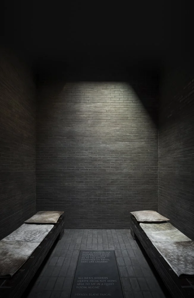Two benches with cushions in a dimly lit, dark brick room with a spotlight on the back wall and engraved text on the floor.
