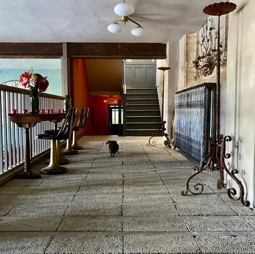 Indoor balcony area with a small dog, floral arrangement on a standing table, staircase, decorative metal radiator, and vintage wall decor.