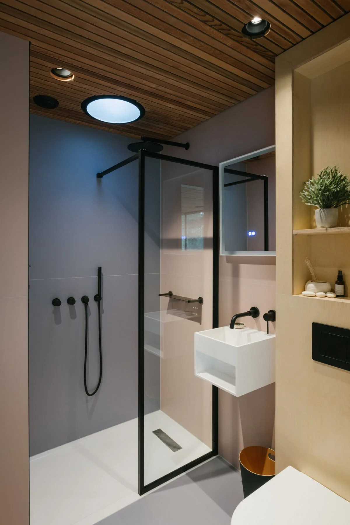 Modern bathroom with walk-in shower, black-framed glass partition, white sink, wooden ceiling, and built-in shelves with plants and toiletries.