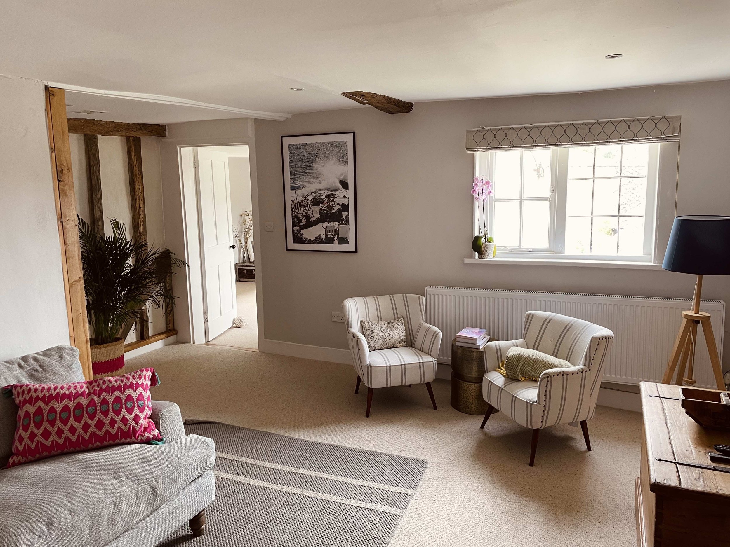 A cozy living room with beige carpet, striped armchairs, a gray sofa with colorful throw pillows, a wooden coffee table, a window with patterned shades, a potted orchid, a black floor lamp, and wooden ceiling beams.