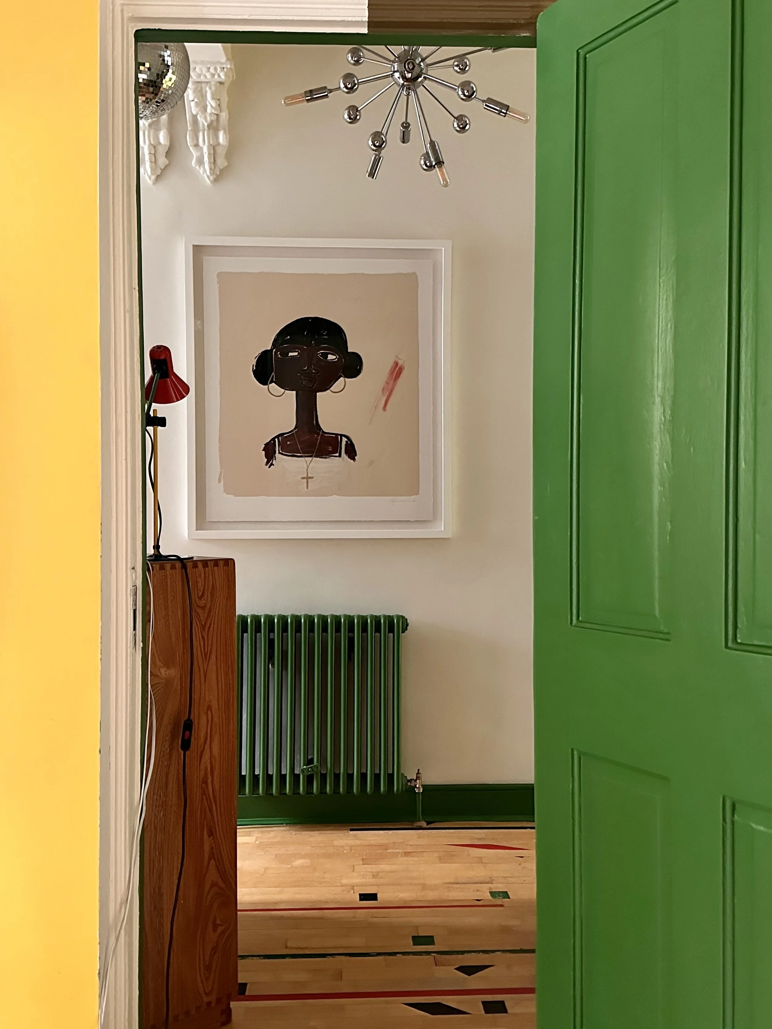View into a room through an open green door, showing framed artwork of a dark-skinned girl with a black bob hairstyle and hoop earrings, a red lamp on a wooden stand, a green radiator, and a wooden floor with geometric inlay.