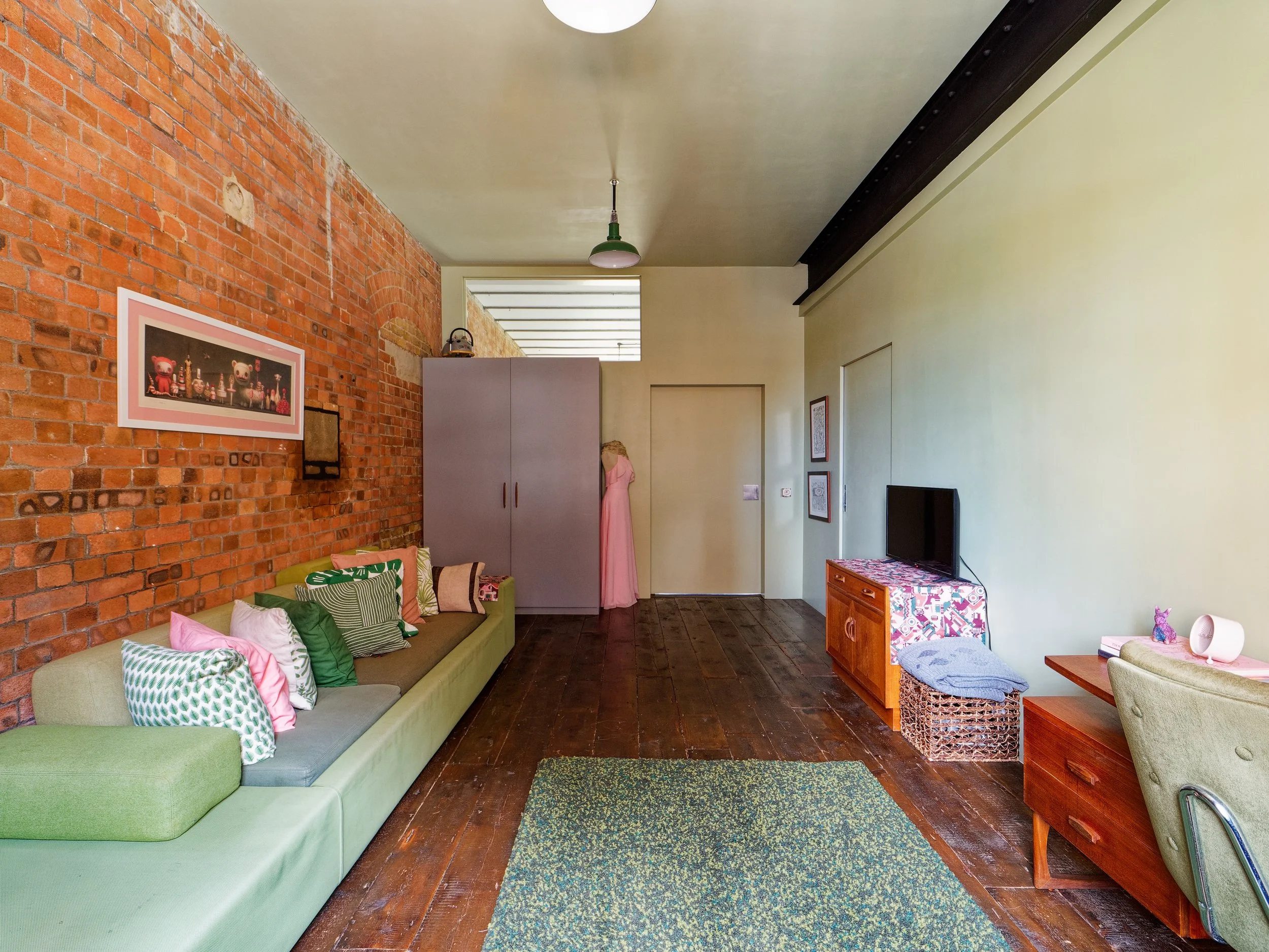 Living room with brick wall, wooden floor, green sofa with pillows, TV on a wooden stand, and a pastel green armchair.