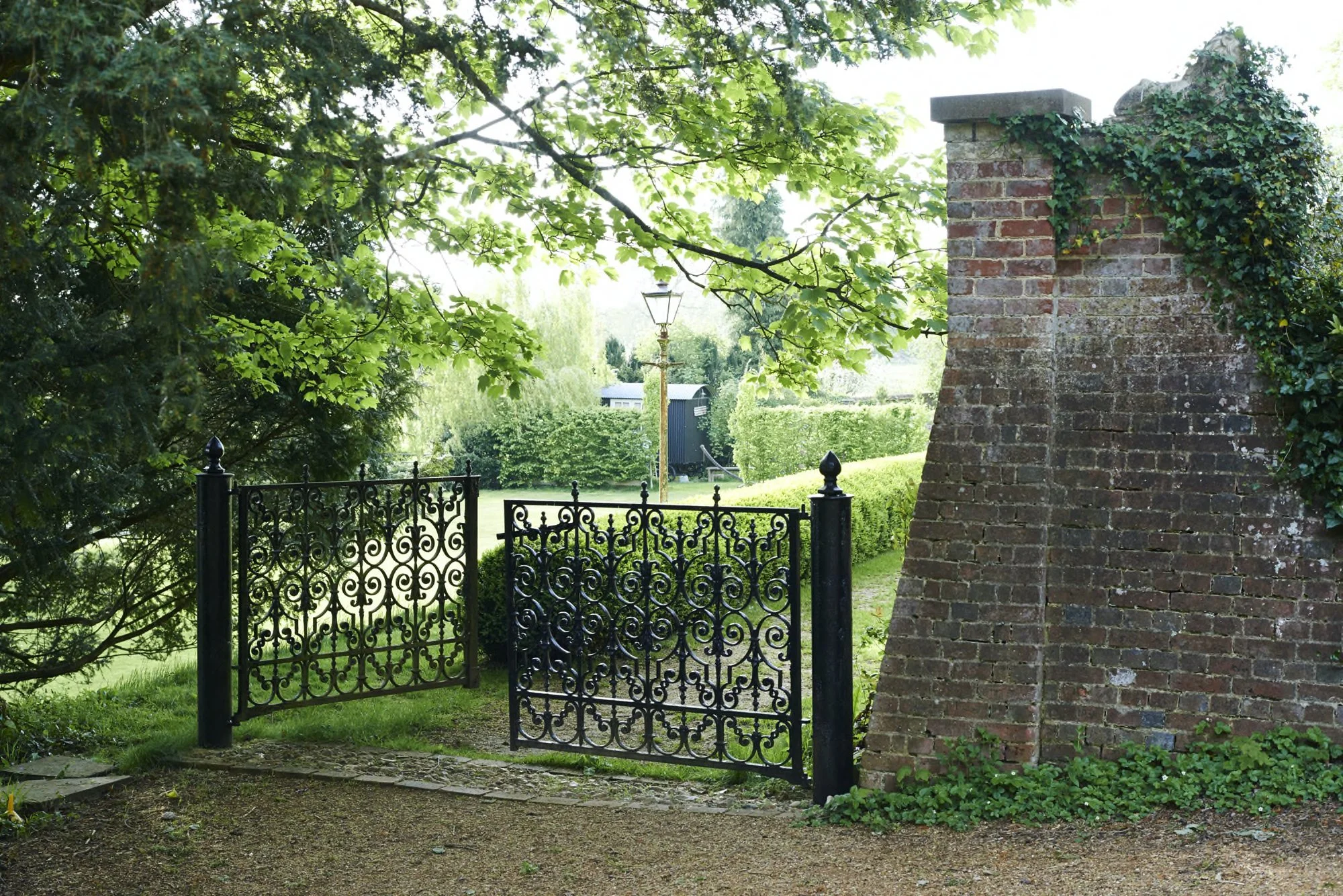 A black wrought iron gate with intricate scrollwork, opening into a lush green garden area, framed by a brick and stone wall on the right and leafy tree branches overhead, with a lawn and hedge in the background.