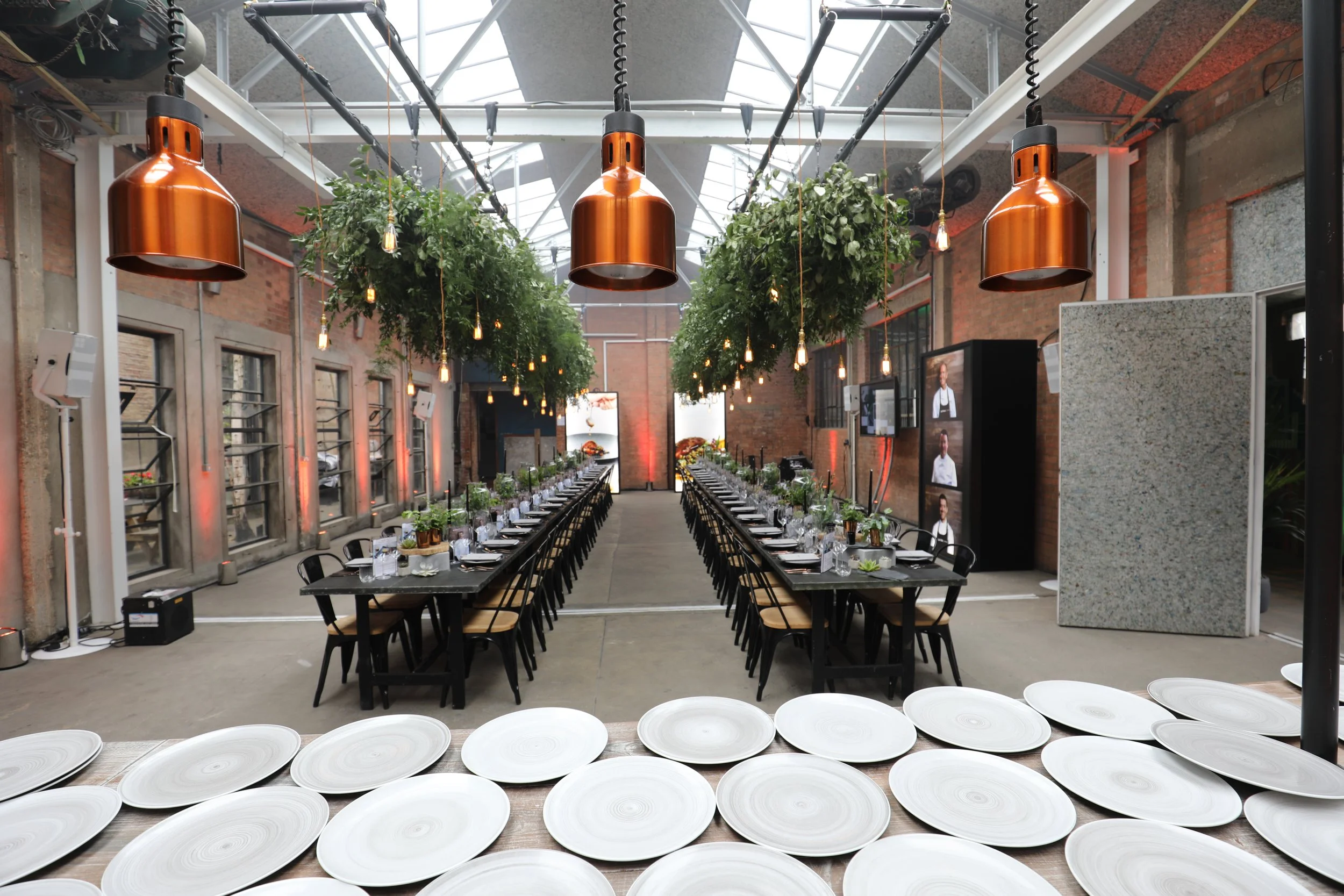 Indoor event space with long dining tables set with plates, glassware, and utensils, hanging plants and copper pendant lights, exposed brick walls, and television screens on the side.