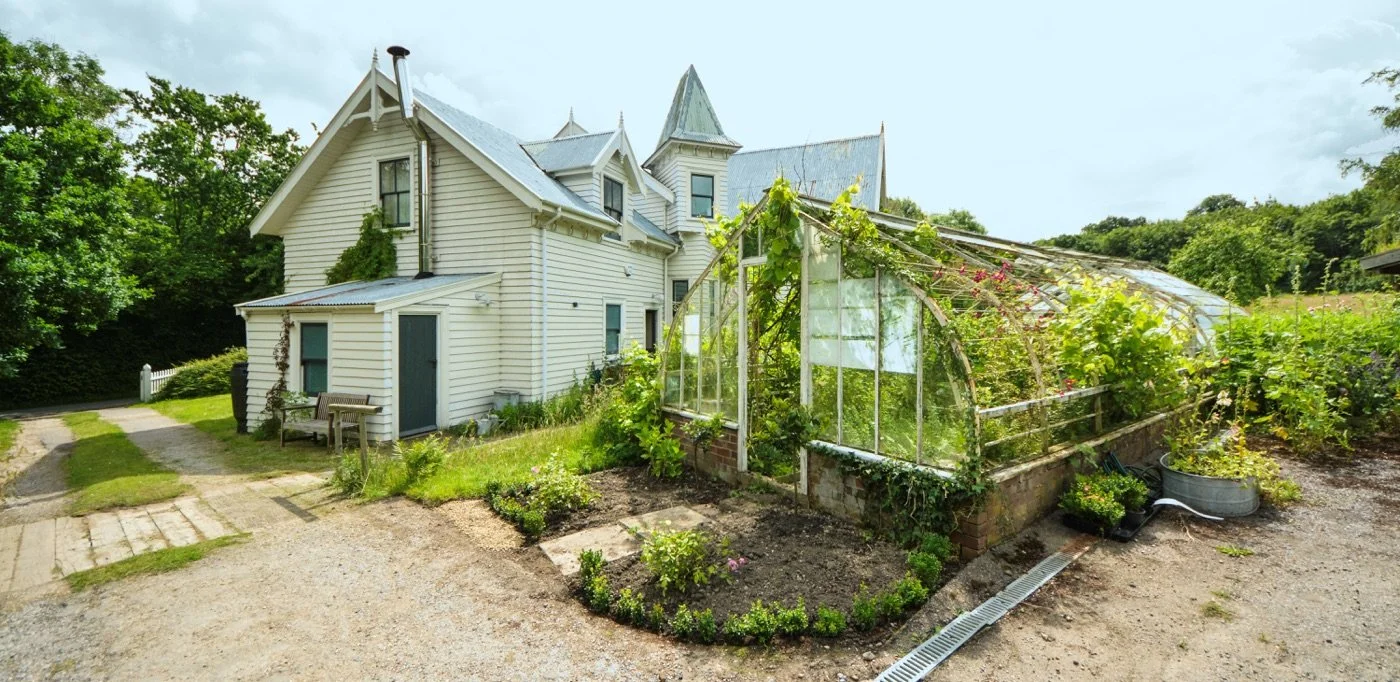 A white house with a green and gray roof, surrounded by trees and a garden with a greenhouse on the right side.