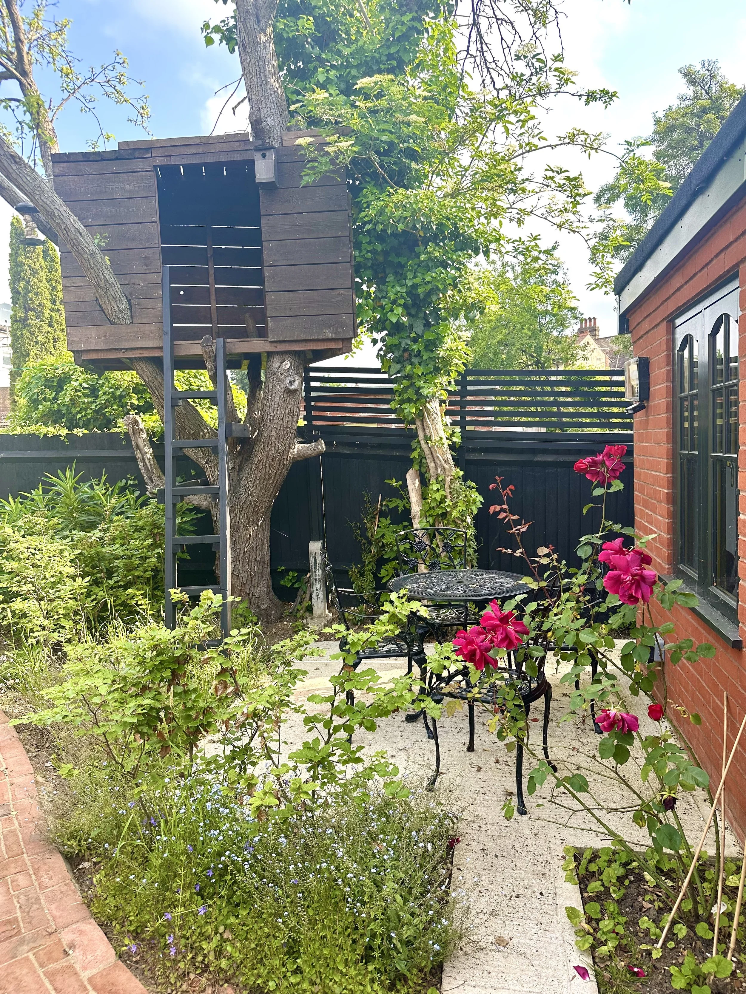 A cozy backyard garden with a treehouse built among the branches of a tree, a small black metal patio table with matching chairs, flowering plants including pink roses, and a brick house wall with black window frames.