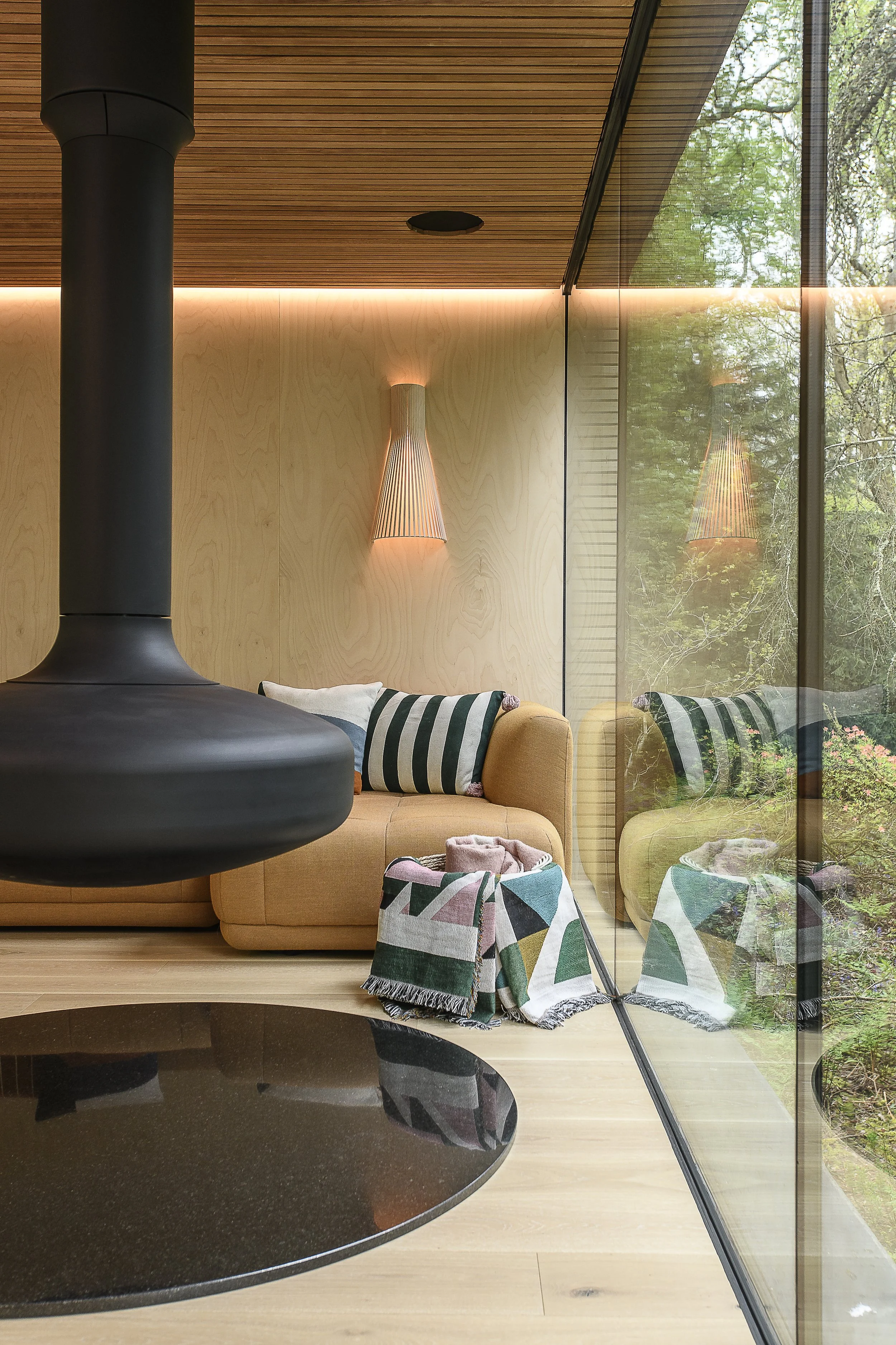 Modern living room with wooden ceiling and walls, a hanging black fireplace, a yellow sofa with striped pillows, a colorful blanket on the floor, a glass wall reflecting trees outside, and wall-mounted light fixtures.