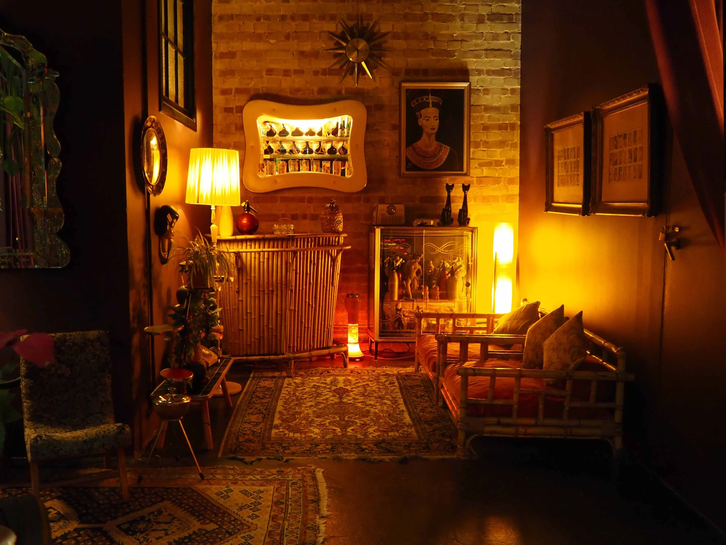 Cozy living room with warm lighting, brick wall, framed artwork of a woman, vintage furniture, rugs, plants, and decorative items.