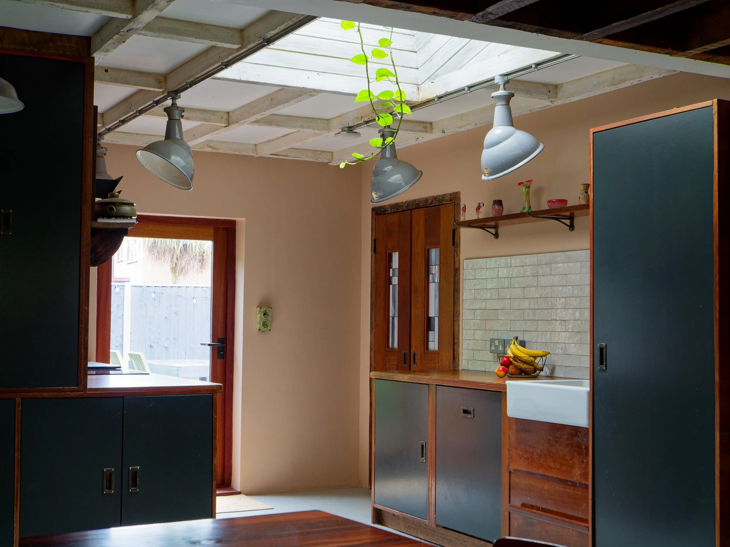 Kitchen with wooden cabinets, a white sink, and a fruit bowl containing bananas, apples, and oranges on the counter. Hanging lights and a skylight above, door leading outside with a view of a patio and fence.