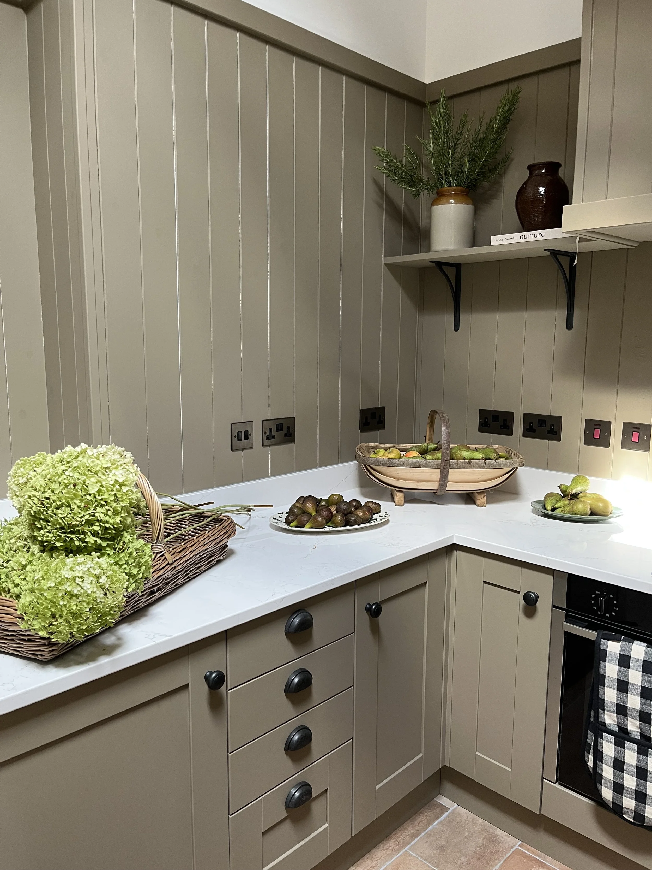 A kitchen corner with beige cabinets, a white marble countertop, and a wall with vertical gray paneling. On the counter, there is a wicker basket with green hydrangeas, a plate with small green fruits, a wooden handle basket with cucumbers, and a pla
