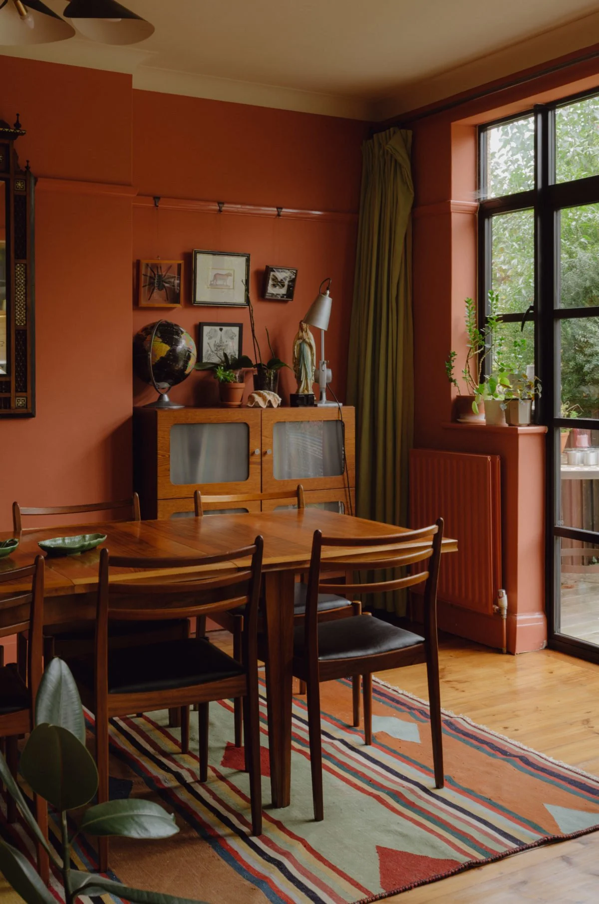 A cozy dining room with a wooden table and six chairs, a colorful striped rug, and a wall of windows with green curtains. There are plants on a cabinet and windowsill, framed pictures, and a globe on the cabinet.